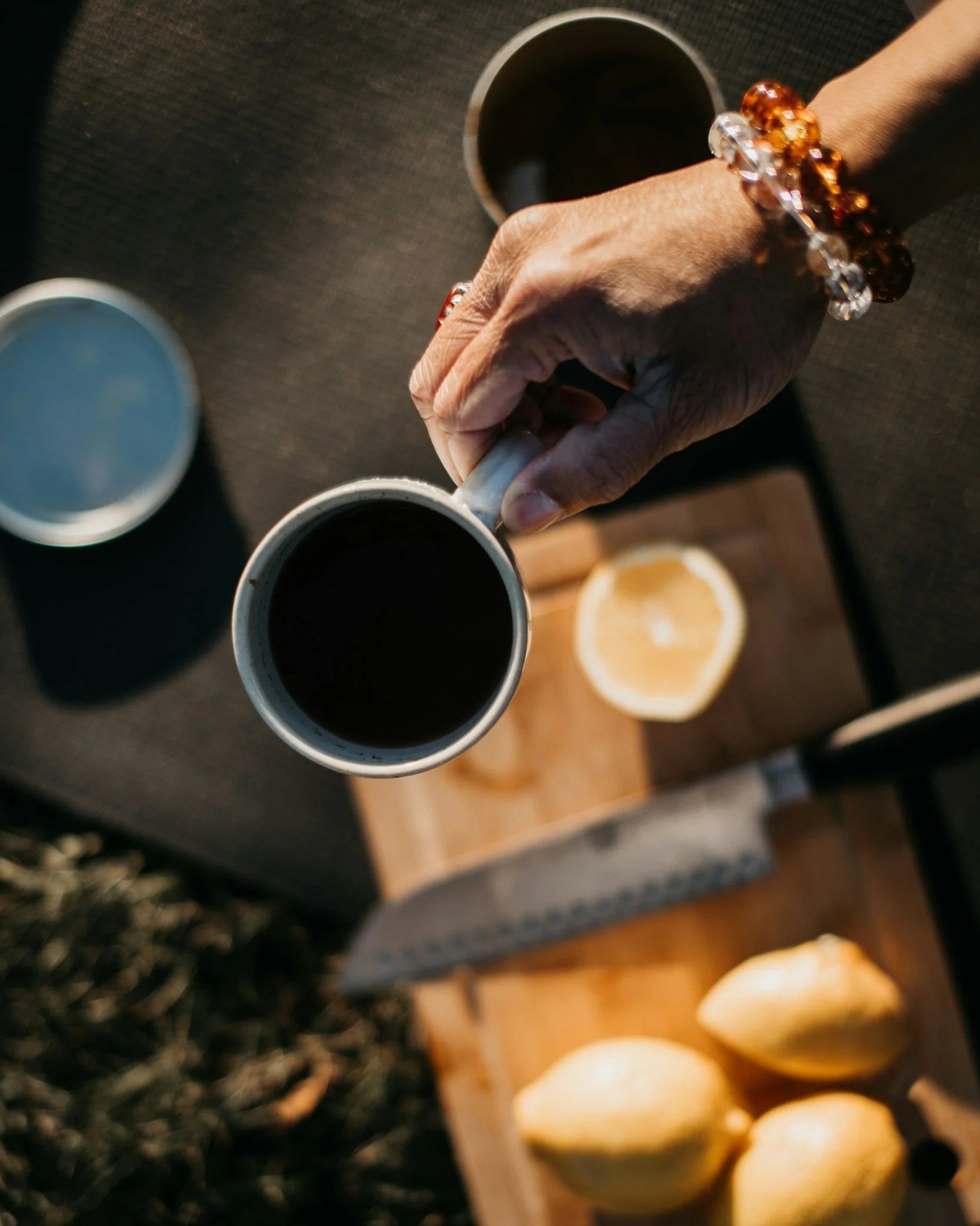 A hand holding a white ceramic mug filled with black coffee, with other coffee cups and food on a wooden table in the background.