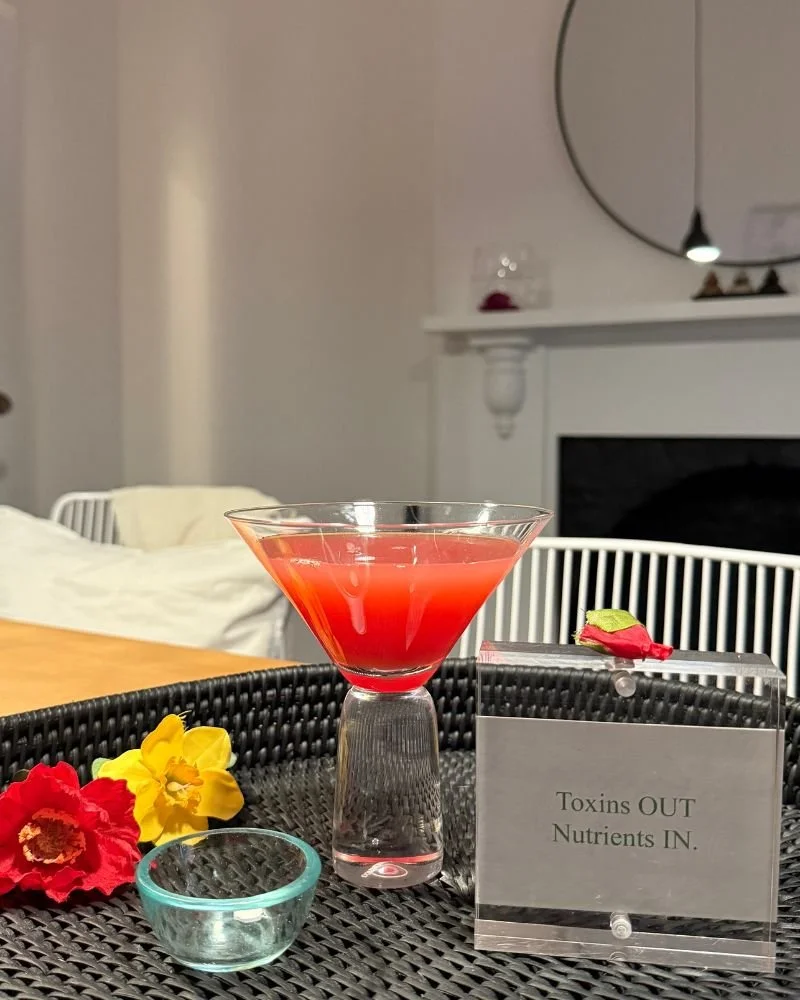 A pink cocktail in a martini glass on a black woven tray. Decorated with red and yellow flowers, and a small glass bowl. A sign reads 'Toxins OUT, Nutrients IN'. Adelaide Colonic Health
