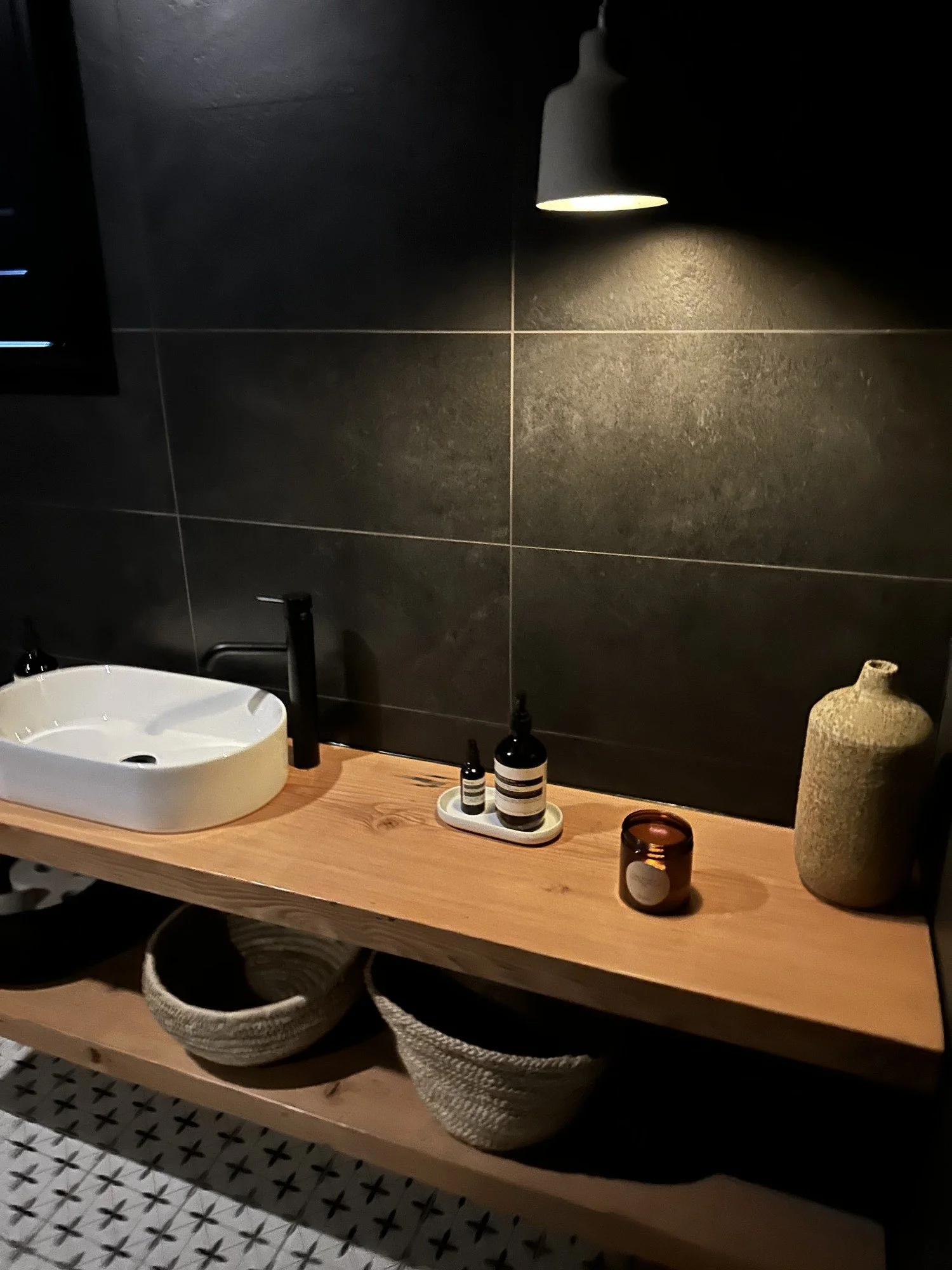 A bathroom countertop with a white oval sink, black faucet, and black soap dispenser. On the wooden shelf below, there are two woven baskets and a dark brown container. The wall behind has large black tiles and a wall-mounted light fixture illuminating part of the wall.