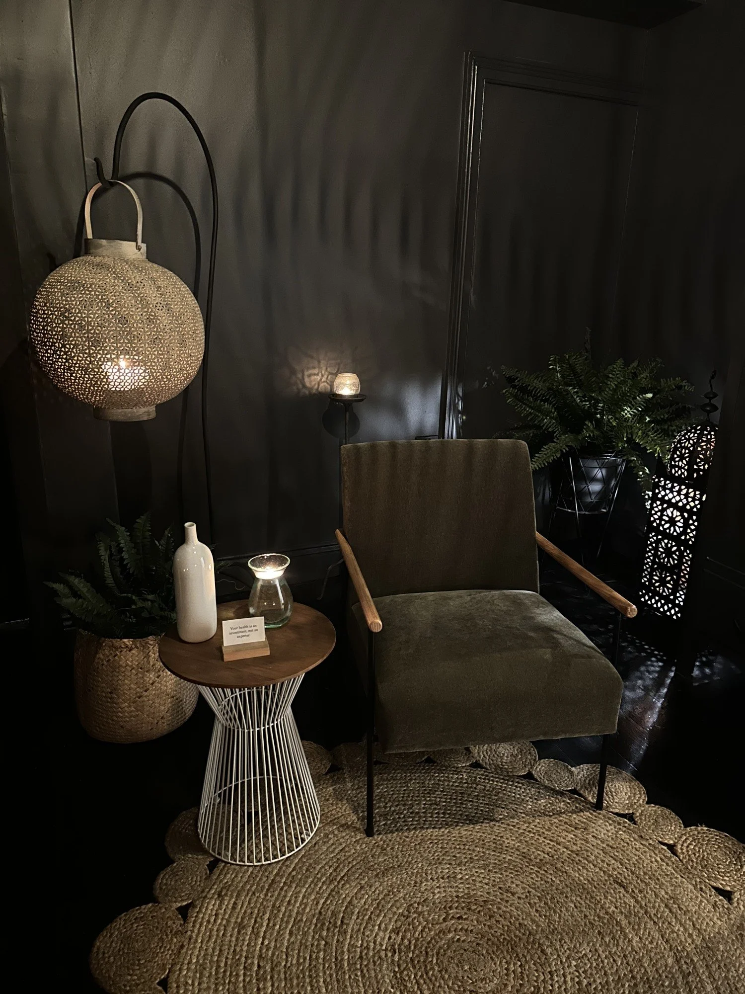 A cozy living room corner featuring a velvet armchair, a round side table with a white wire base, a white vase, a small glass, and a note card, illuminated by a patterned hanging lamp and a small table lamp, with plants and decorative panels in the background.
