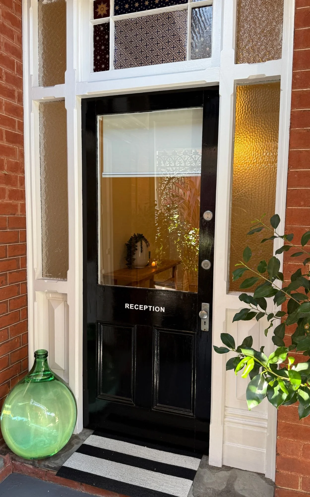 Black reception door with glass window, white trim, and the word 'RECEPTION' in white letters, surrounded by brick and textured glass side panels, with indoor greenery visible through the window and a striped black and white rug outside.