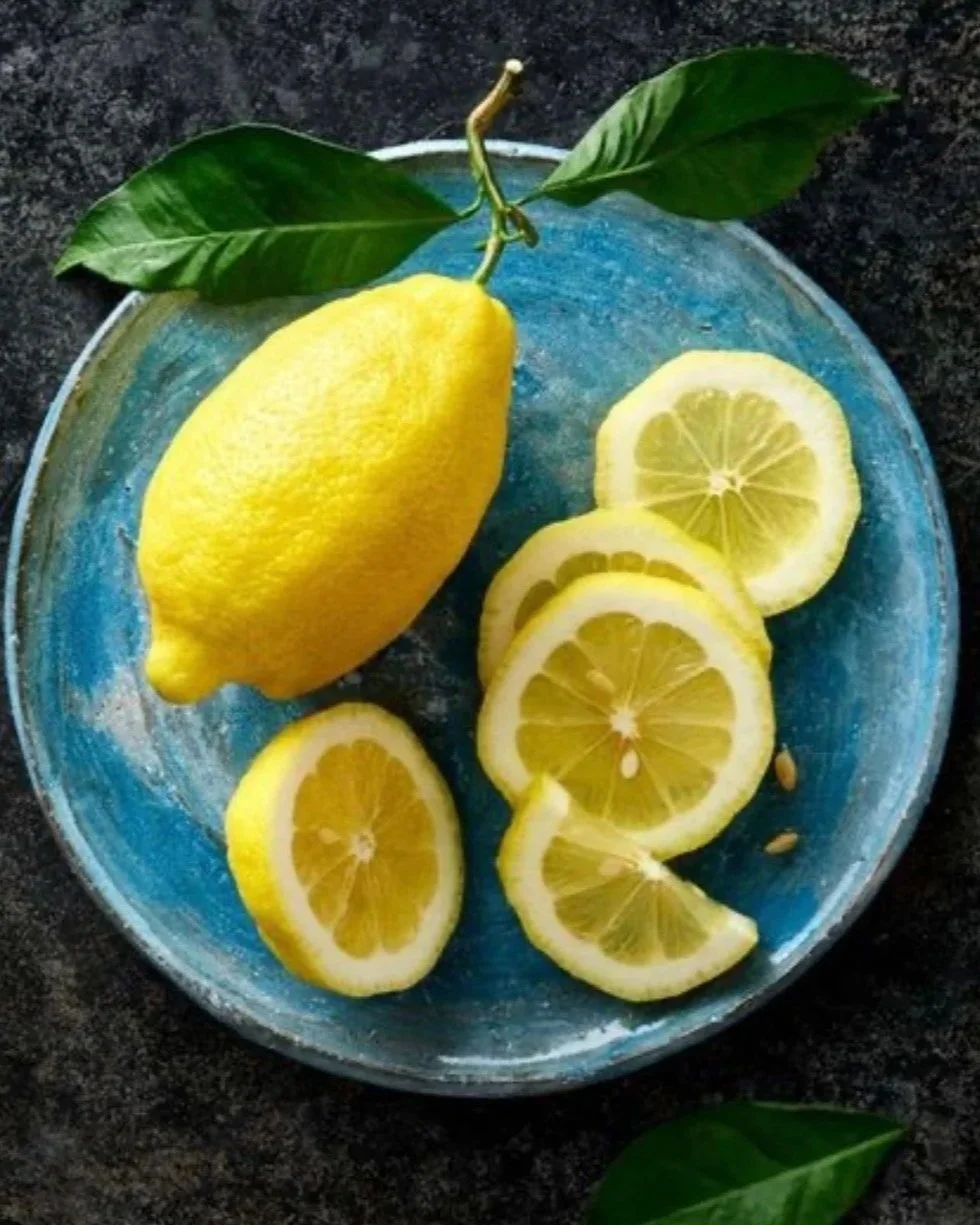 Whole yellow lemon with green leaves and several sliced lemon rounds on a blue plate on a dark surface.