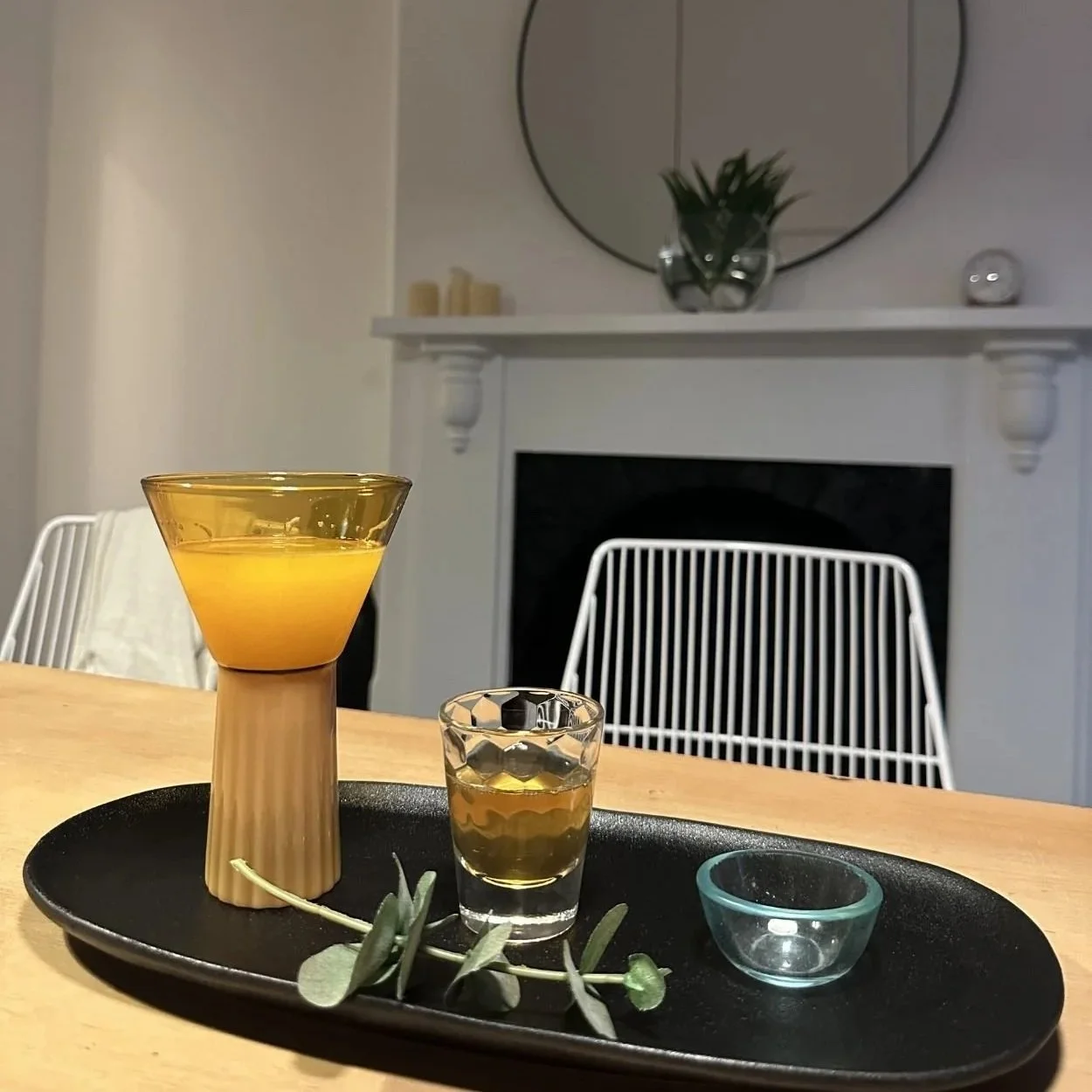 A black tray on a wooden table holding two glasses and decorative items, with a living room background featuring a white fireplace mantel, mirror, plant, candles, and a white chair.