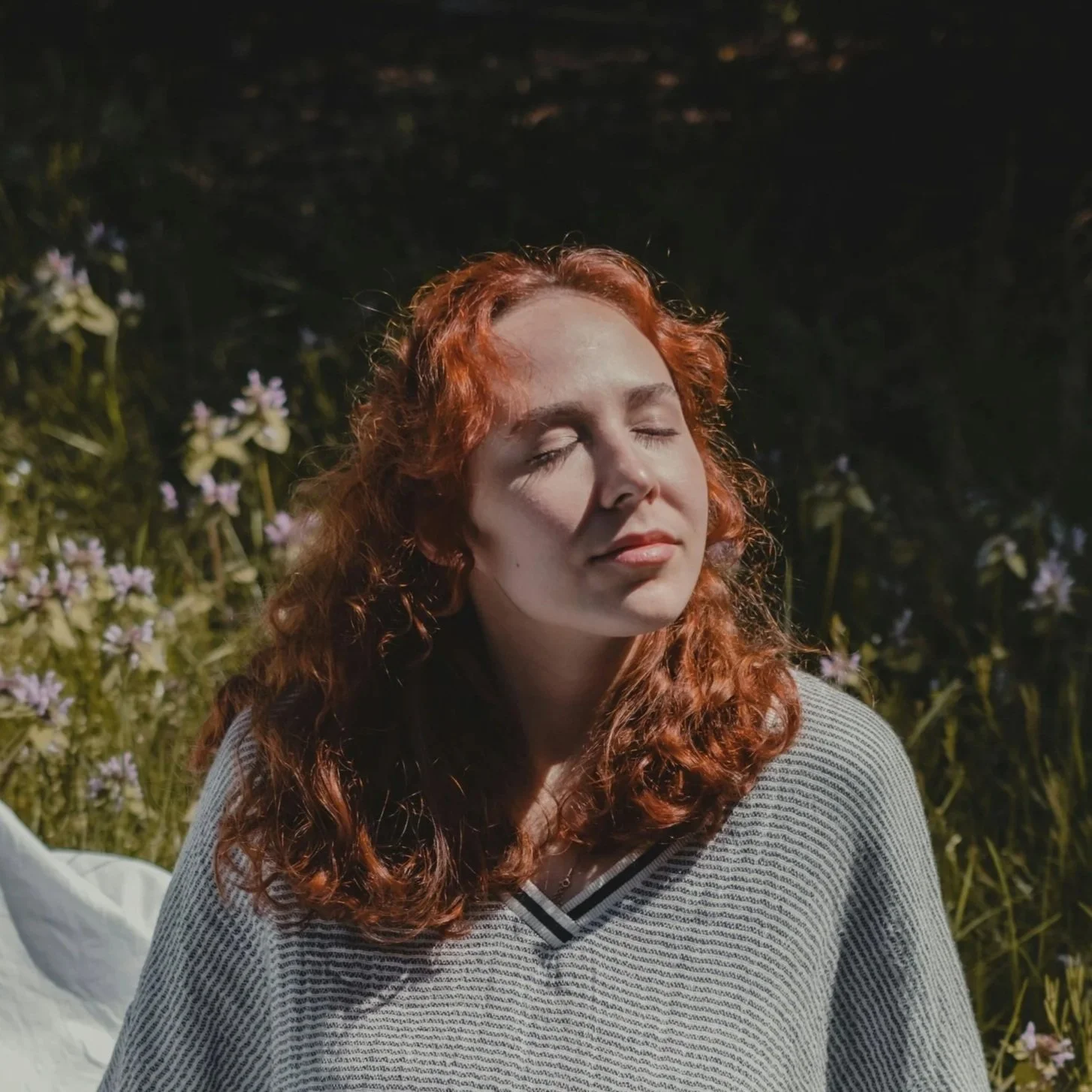 A woman with red curly hair, eyes closed, outdoors at night with flowers and greenery in the background.