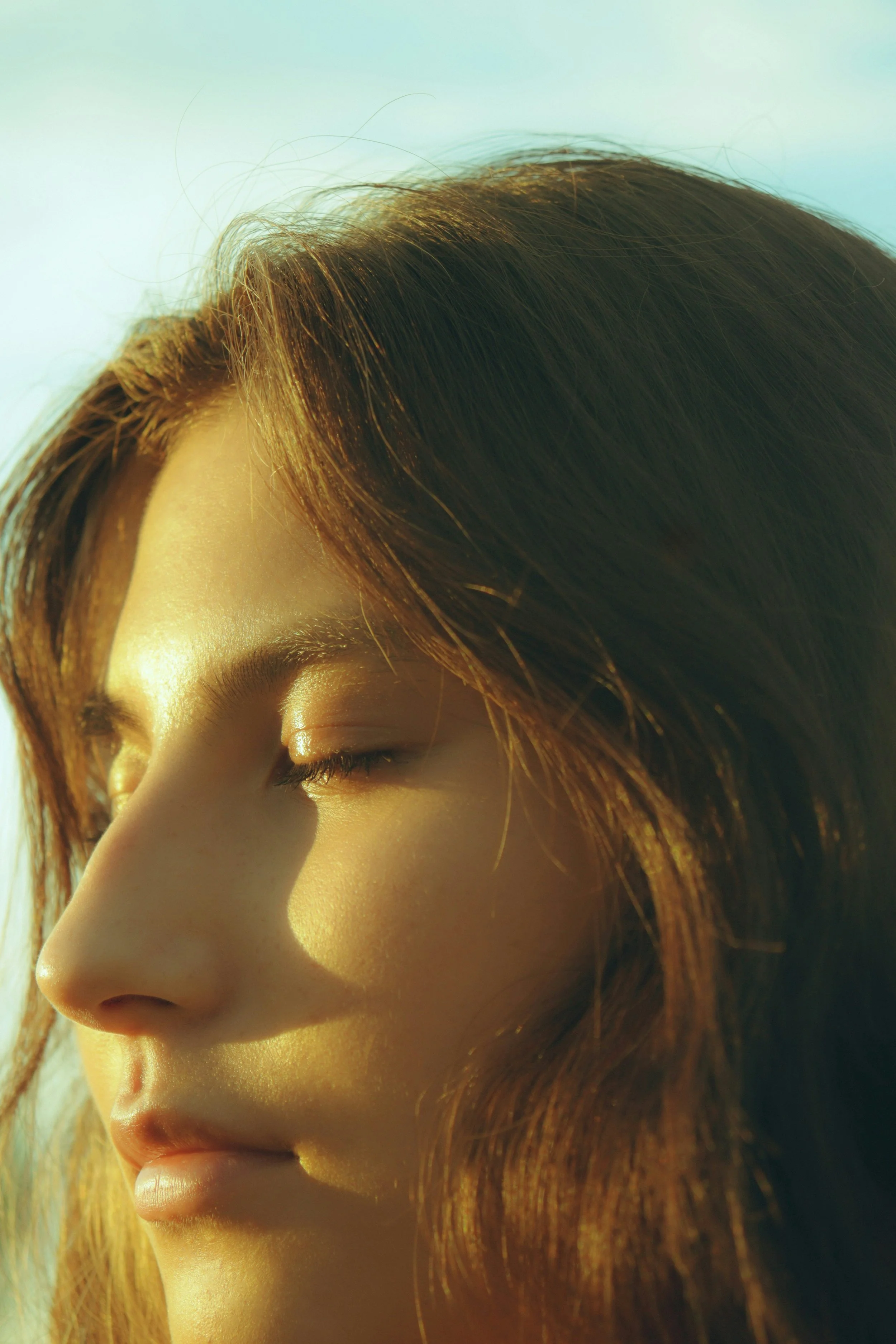 Close-up of a woman's face with closed eyes and reddish-brown hair, illuminated by warm sunlight.