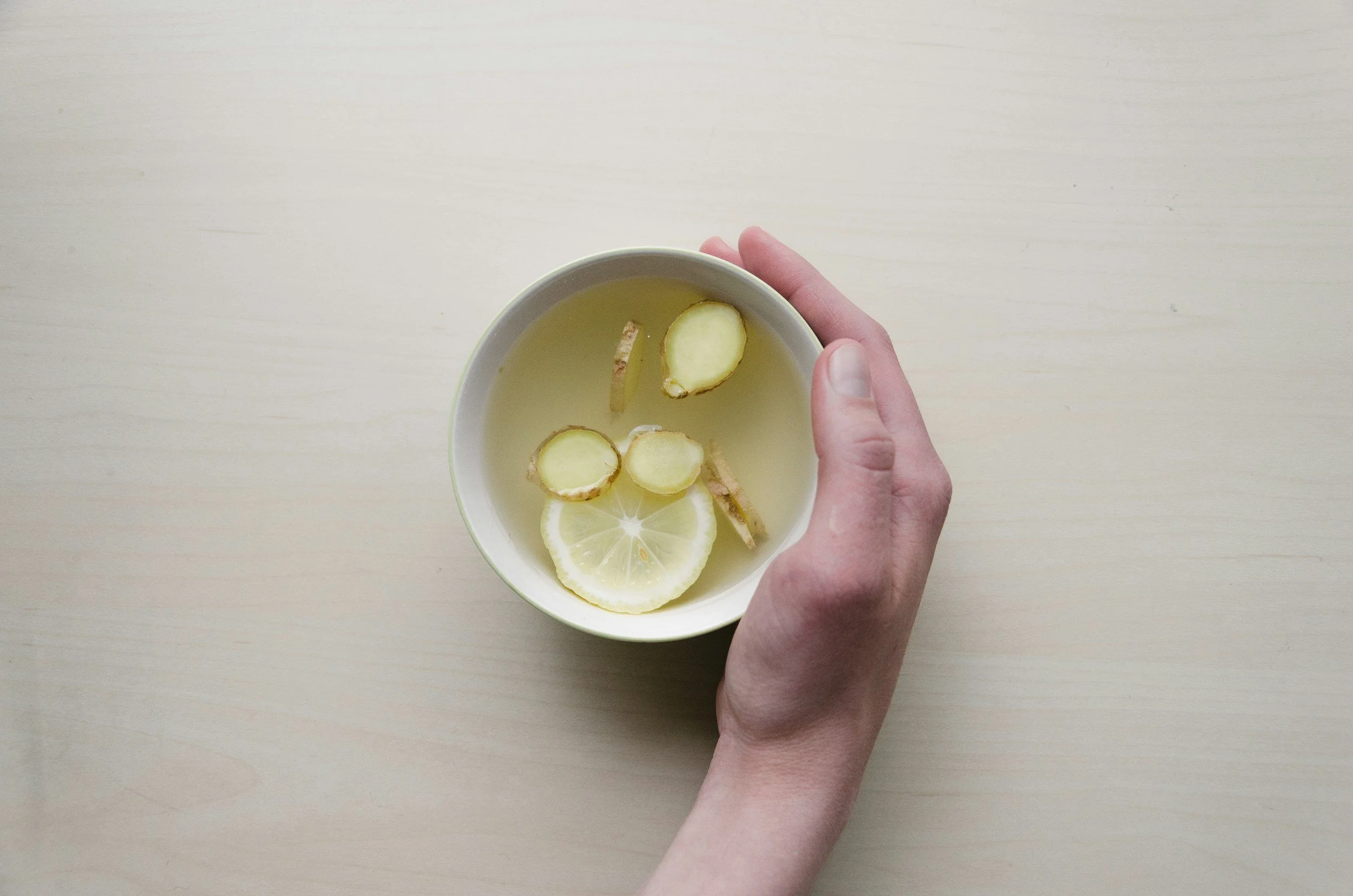 A person holding a white bowl with slices of lemon, ginger, and cucumber in a light-colored liquid.
