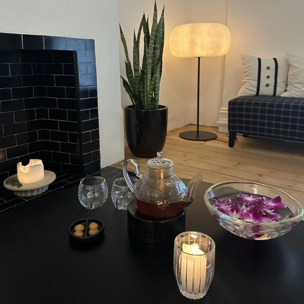 Living room with a black brick fireplace, a potted snake plant, a tall floor lamp with a round shade, a plaid sofa with pillows, a teapot with tea, a bowl of purple orchids, and candles on a black table.