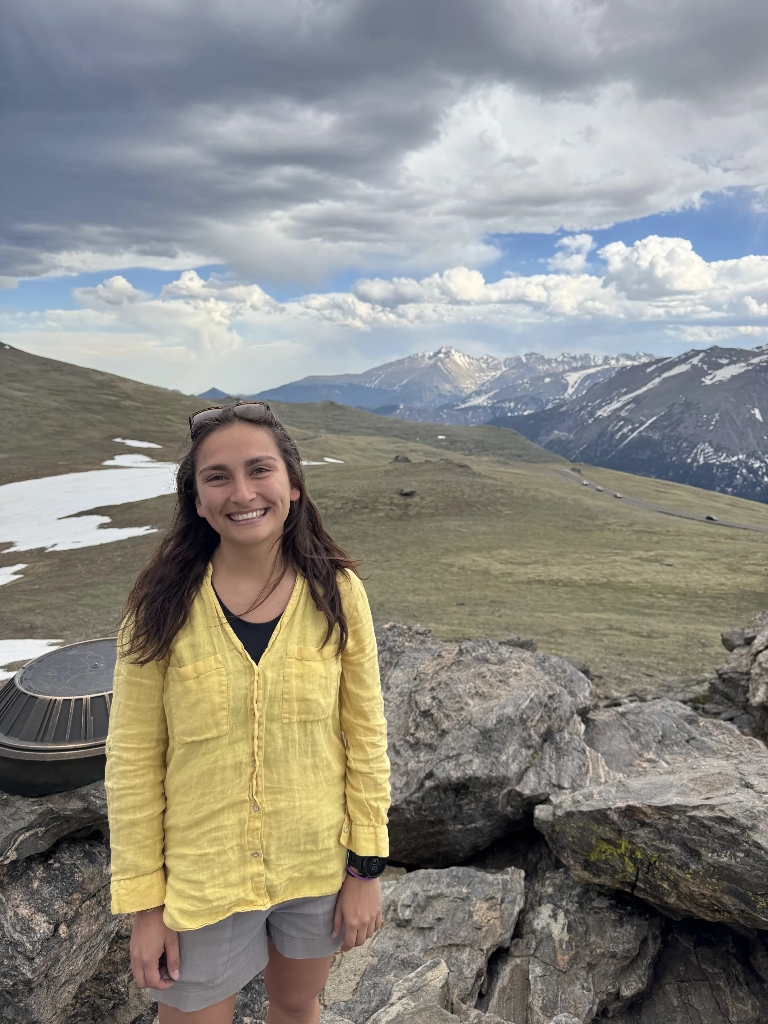 A young woman smiling outdoors in a mountainous landscape with snow-capped peaks, grassy slopes, and cloudy sky, standing on rocky terrain.