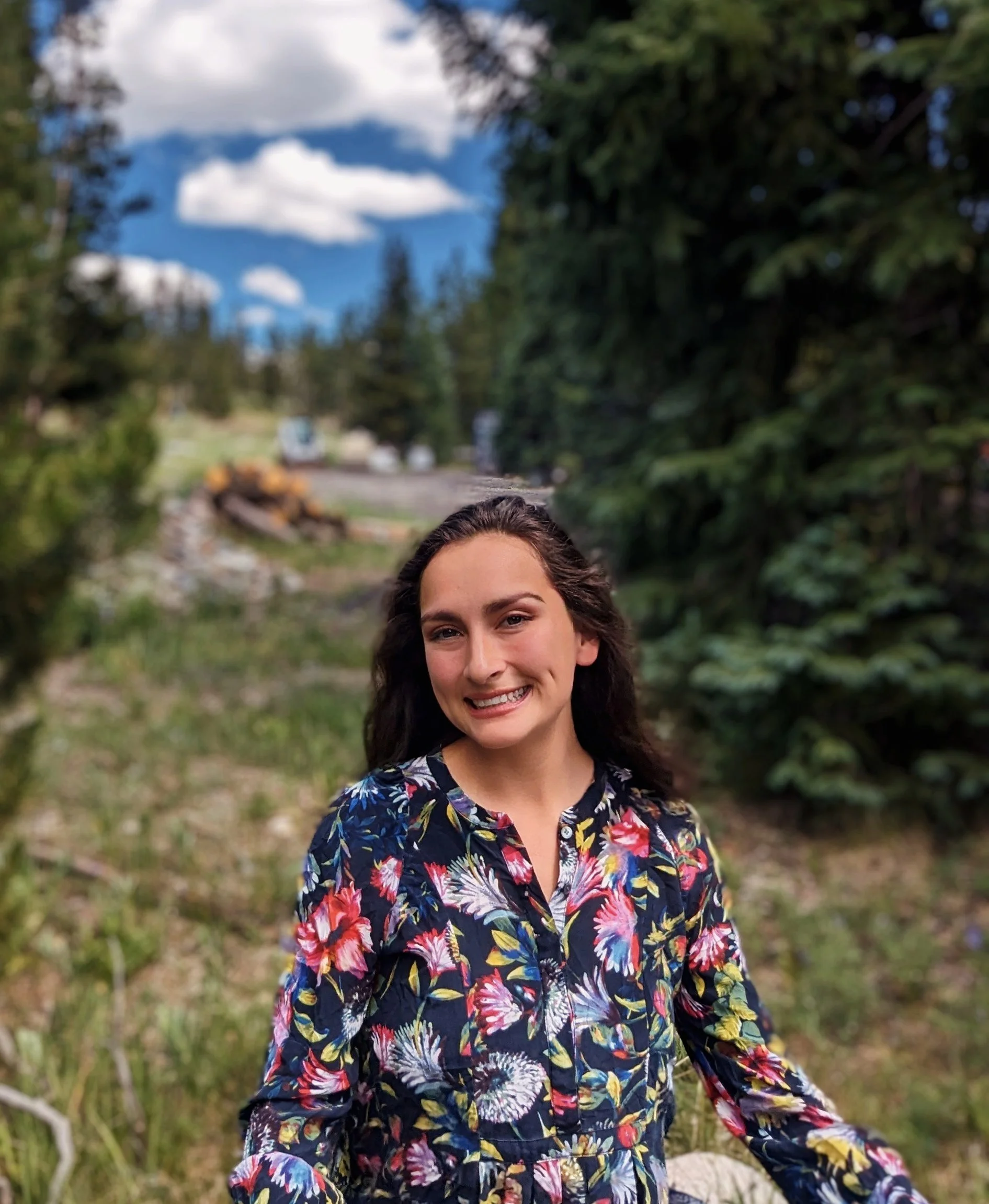 Smiling woman with long dark hair wearing a colorful floral blouse outdoors with trees and blue sky with clouds in the background in Summit County.