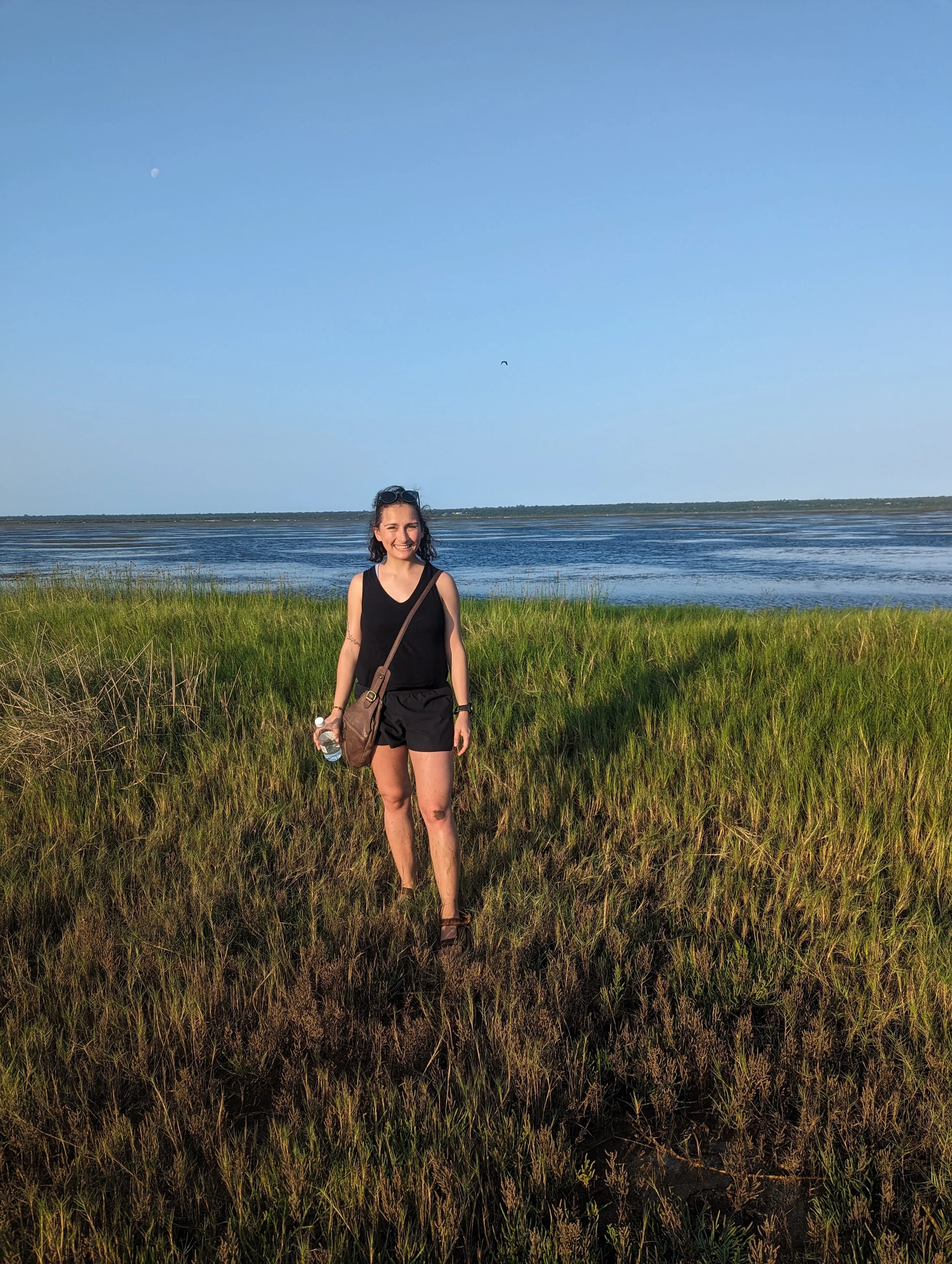 A woman standing in a grassy field near water, smiling, with the moon and a bird in the sky behind her.