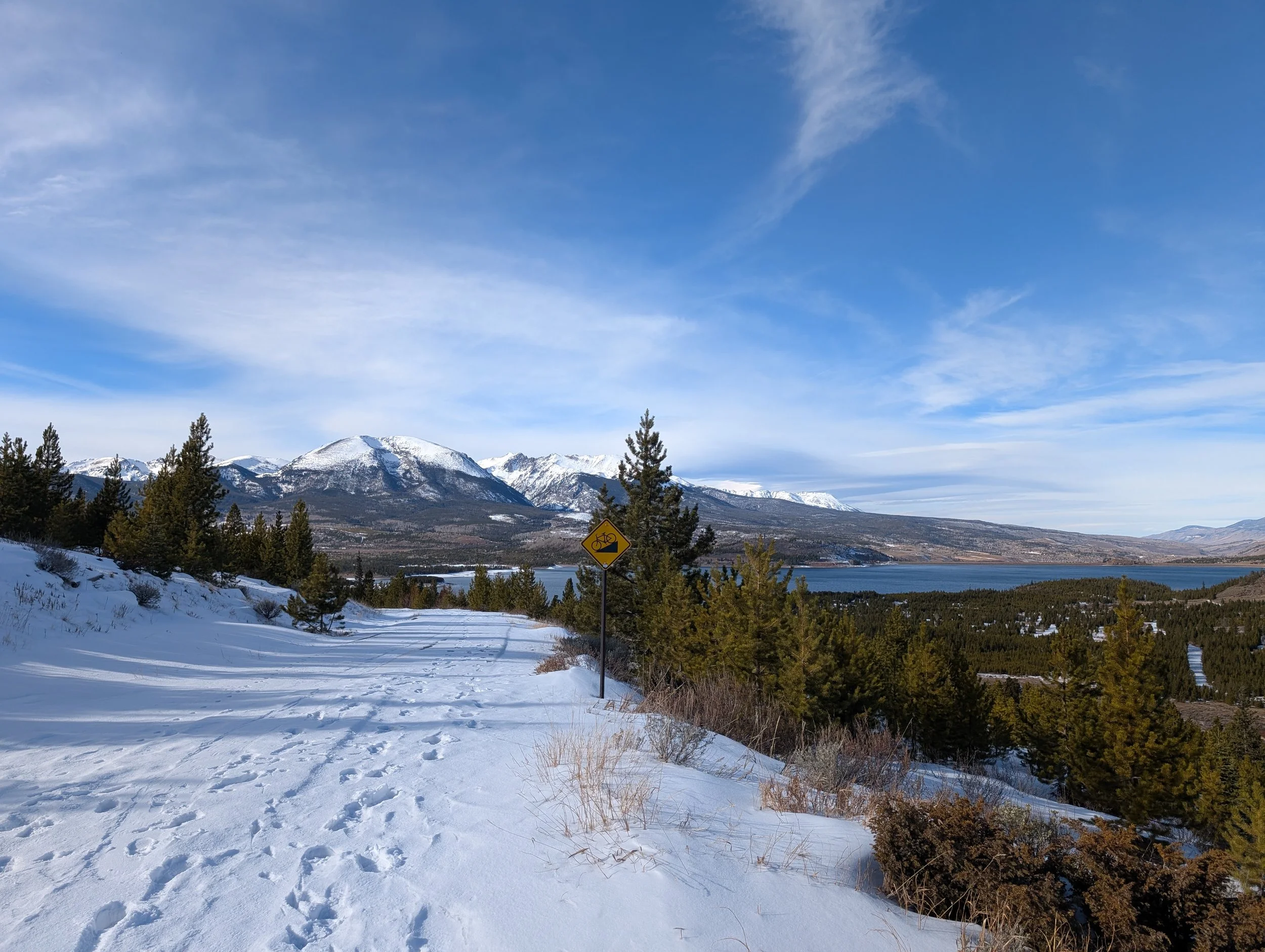 Snow-covered ground with tire tracks and footprints, pine trees, a river, mountains with snow, and a partly cloudy sky.