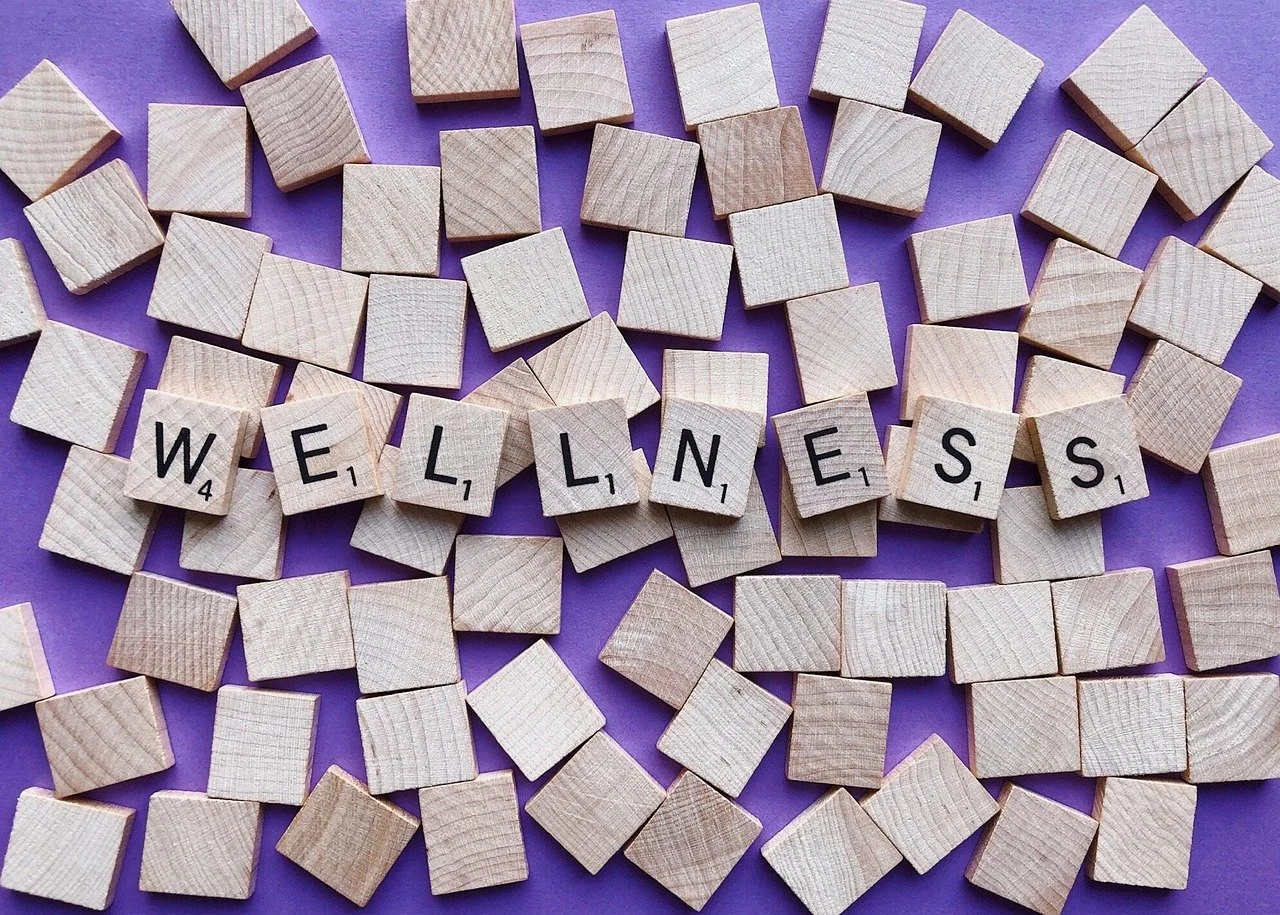 Wooden Scrabble tiles spelling out 'WELLNESS' on a purple background with scattered wooden blocks around.