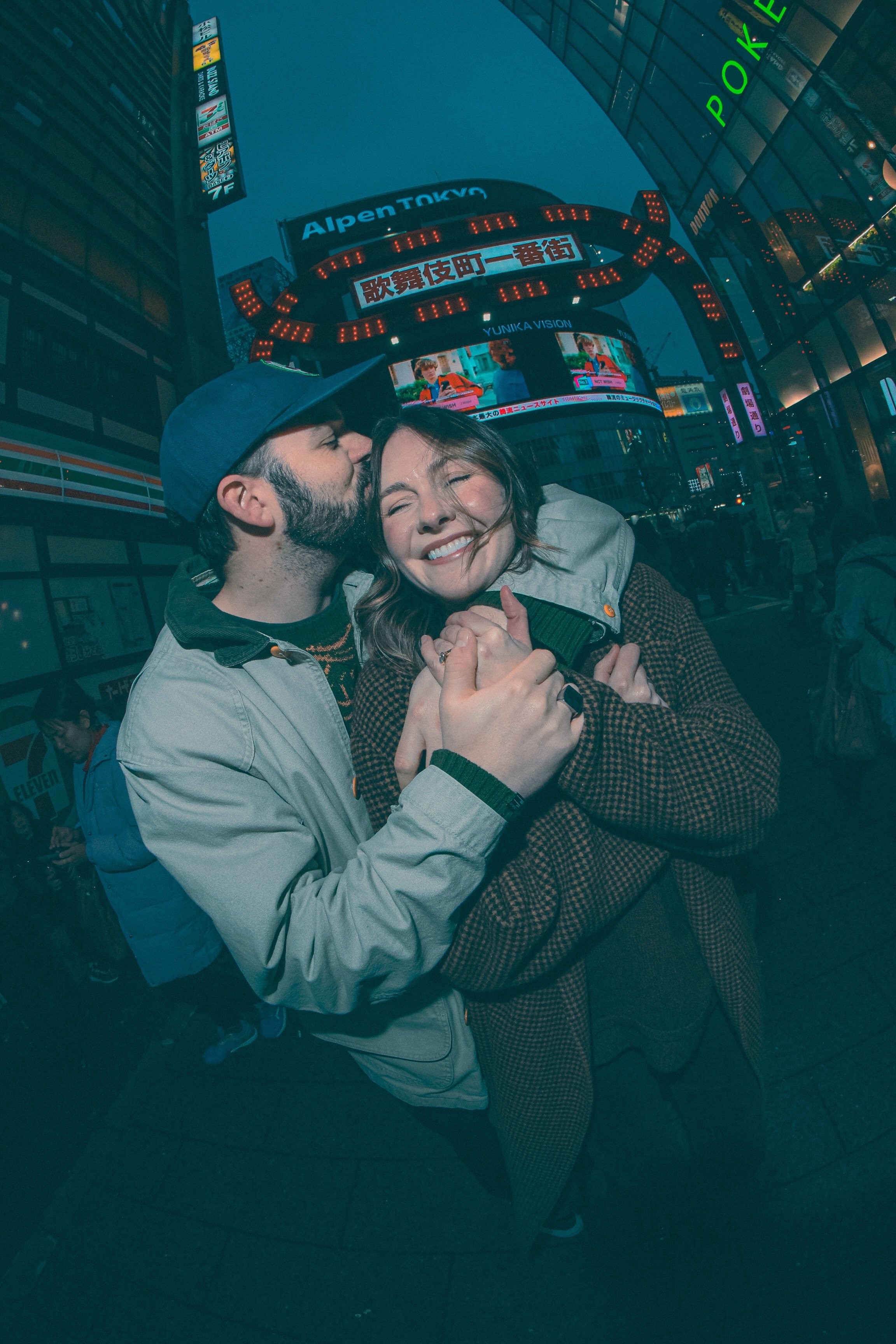 A young man and woman sharing a kiss in Times Square, New York City, at night, with bright electronic billboards and signs in the background.