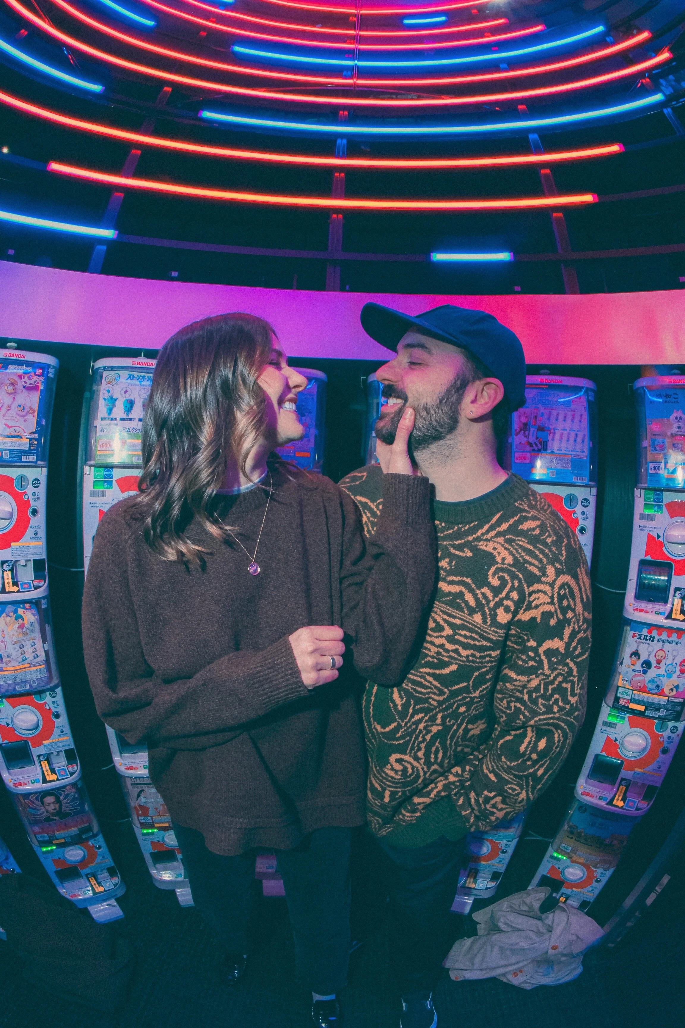 A young woman and a young man are smiling and laughing on a brightly lit arcade with colorful neon lights and claw machines in the background.