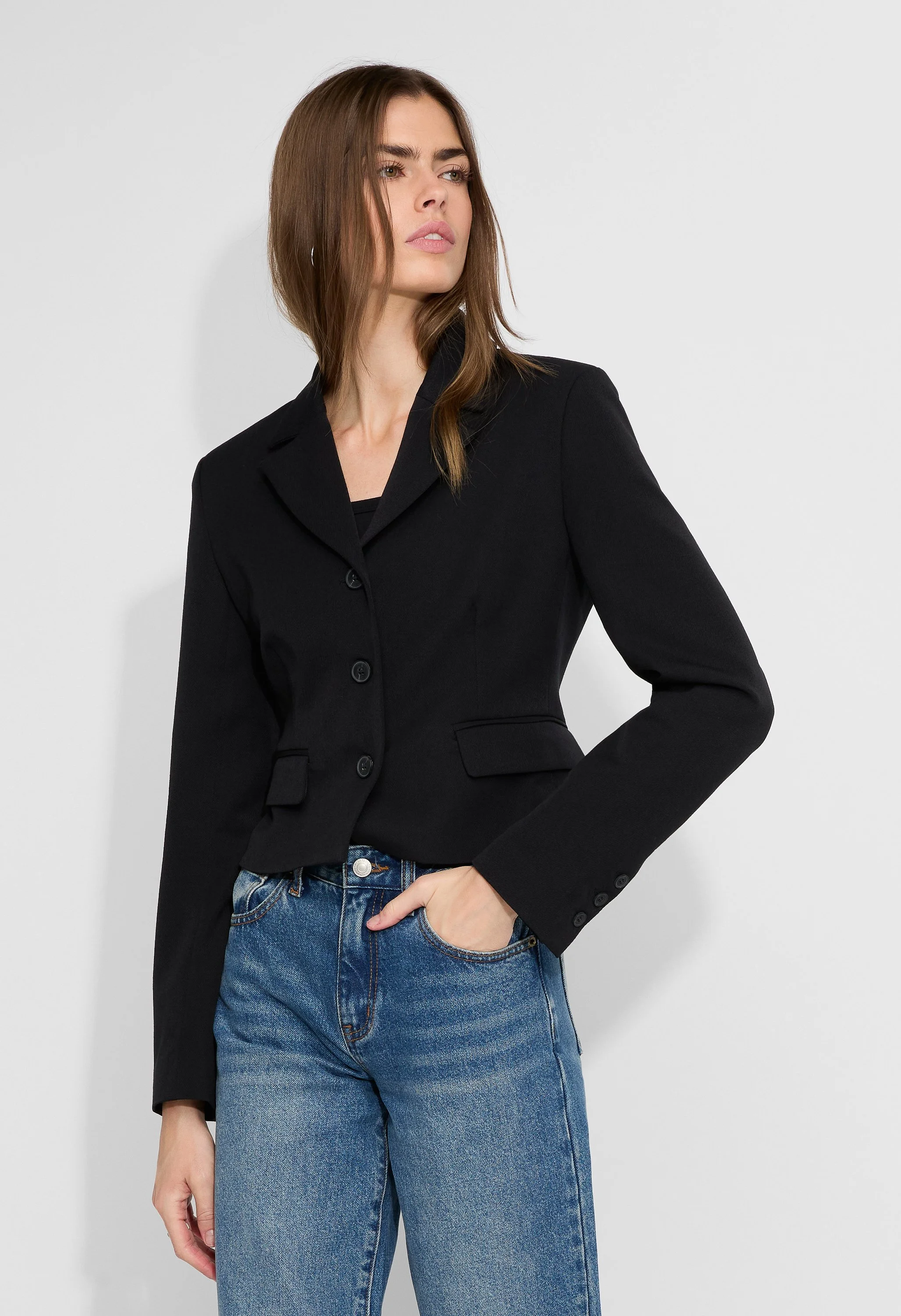 A woman with shoulder-length brown hair wearing a black blazer and blue jeans stands against a plain white wall. She has a neutral expression and looks to her left.