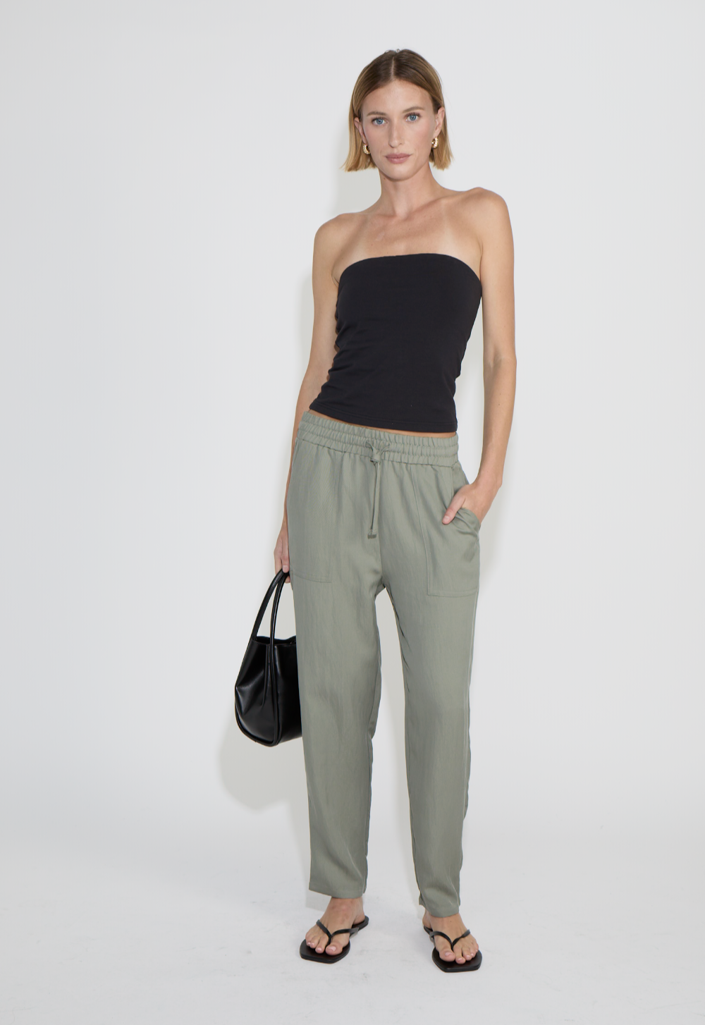 A woman standing against a plain light gray background, wearing a strapless black top, olive green loose pants with an elastic waistband, black sandals, carrying a black handbag, standing against a white background in an e-commerce studio.
