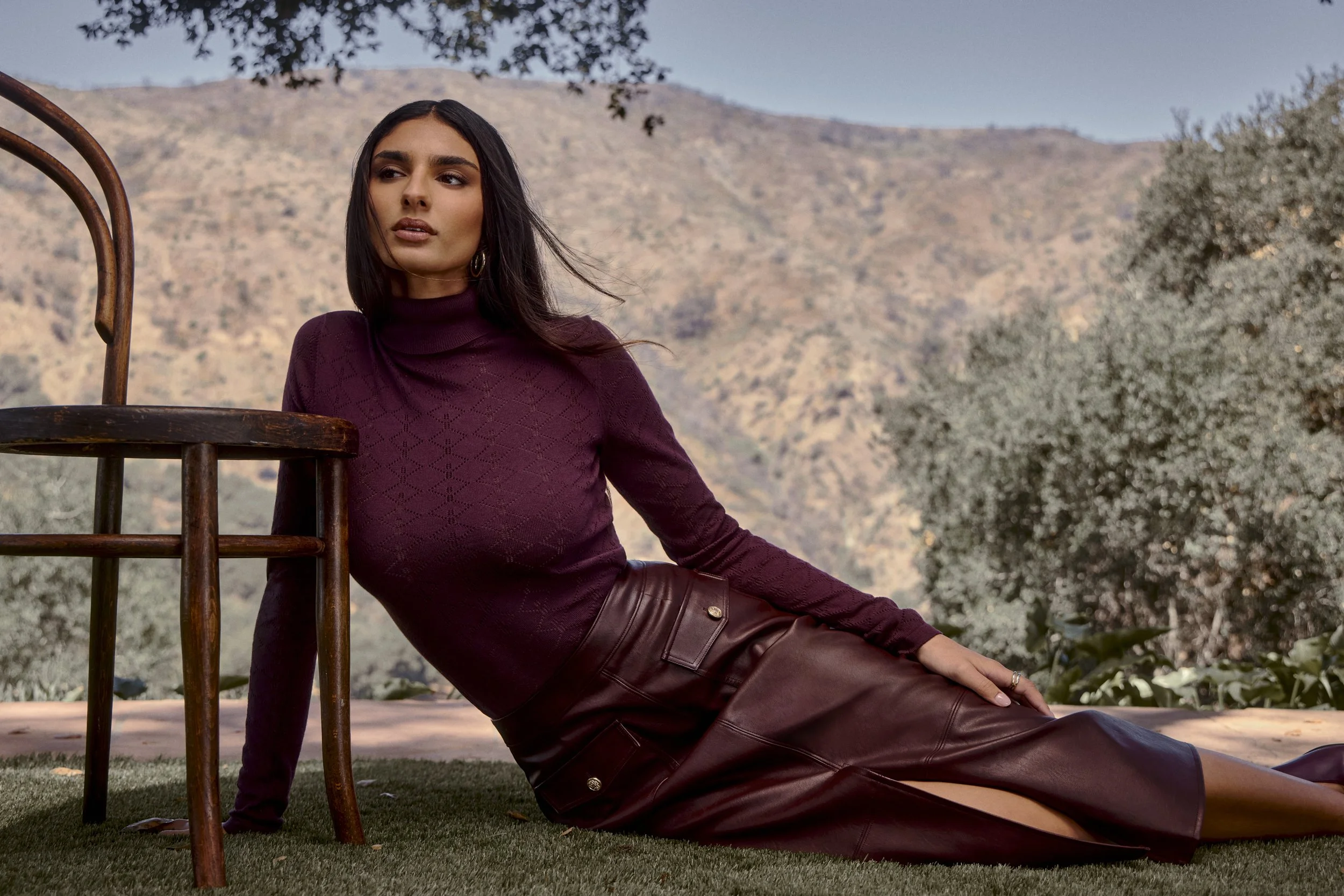 A woman with dark hair, wearing a maroon turtleneck sweater and leather skirt, posing outdoors near a wooden chair with mountains and trees in the background.