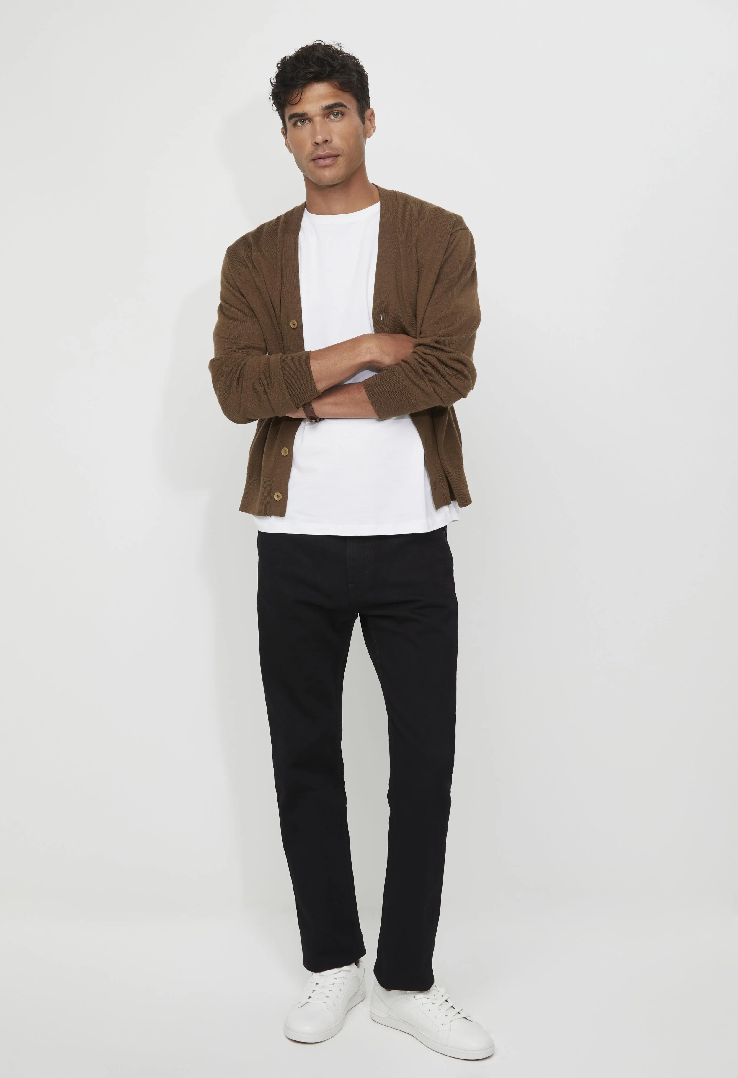 Young man standing against a plain white wall, wearing a brown cardigan over a white T-shirt, black pants, and white sneakers, with arms crossed and looking at the camera.