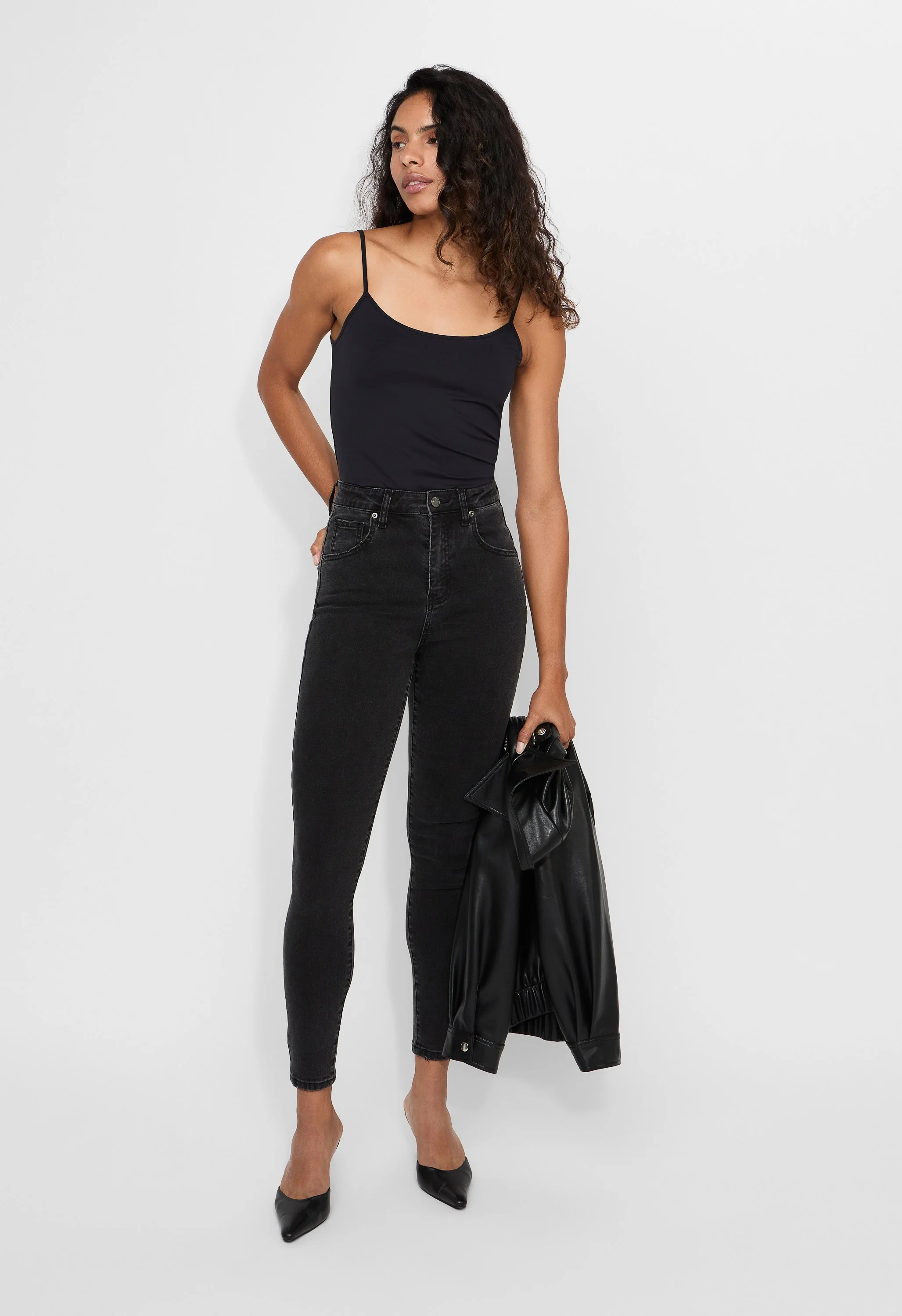 A woman with curly hair and tan skin wearing a black tank top, high-waisted black jeans, black heels, holding a black leather jacket and a clutch, standing against a plain white background in an e-commerce studio.