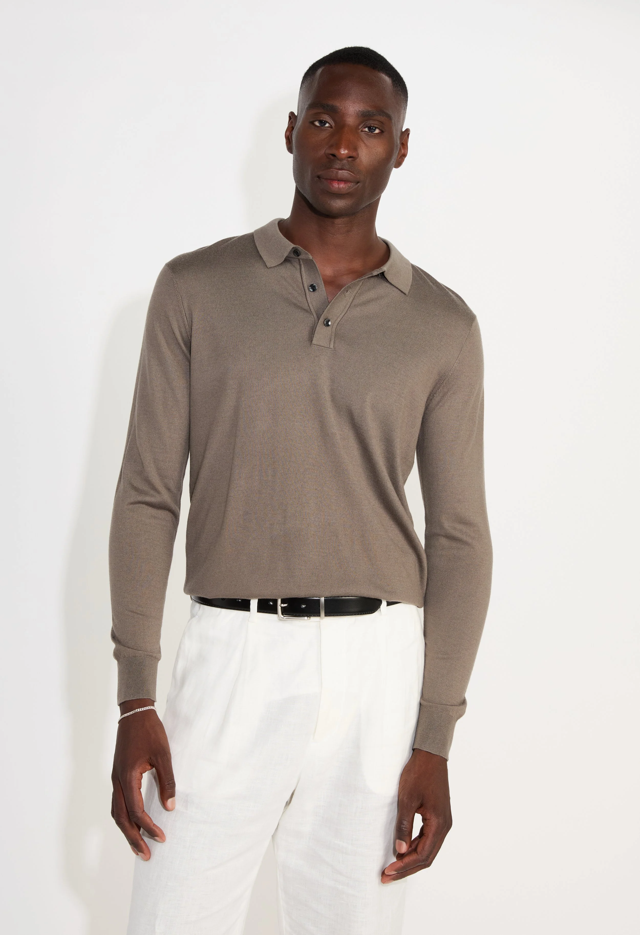 Man wearing a taupe polo shirt and white pants standing against a white background.