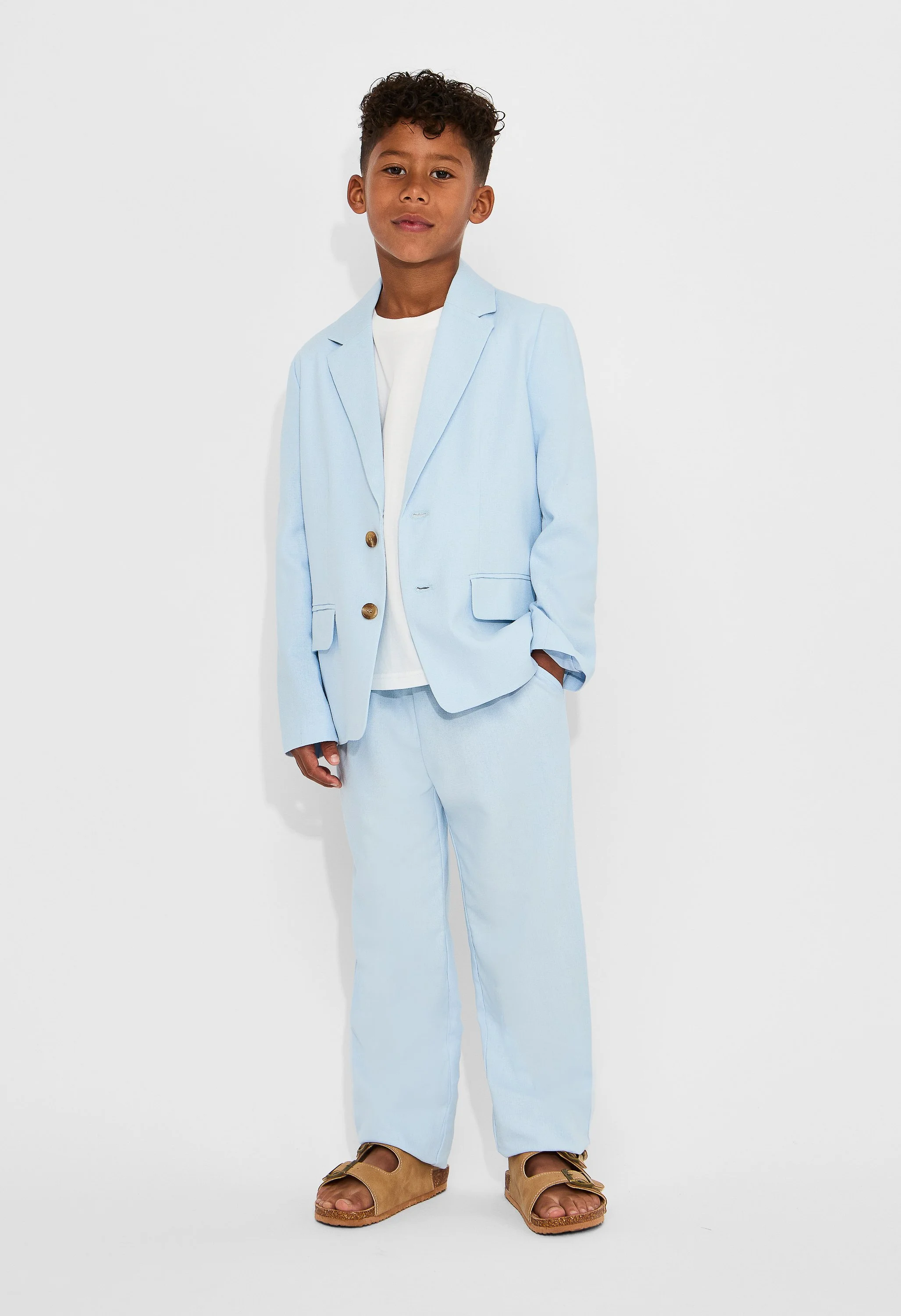 A young boy with curly hair, wearing a light blue blazer and matching pants, white T-shirt, brown sandals, standing against a plain white background, looking confidently at the camera.