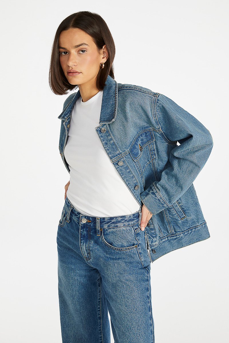 A woman with shoulder-length dark hair wearing a denim jacket over a white t-shirt and matching denim jeans, standing against a plain white background.