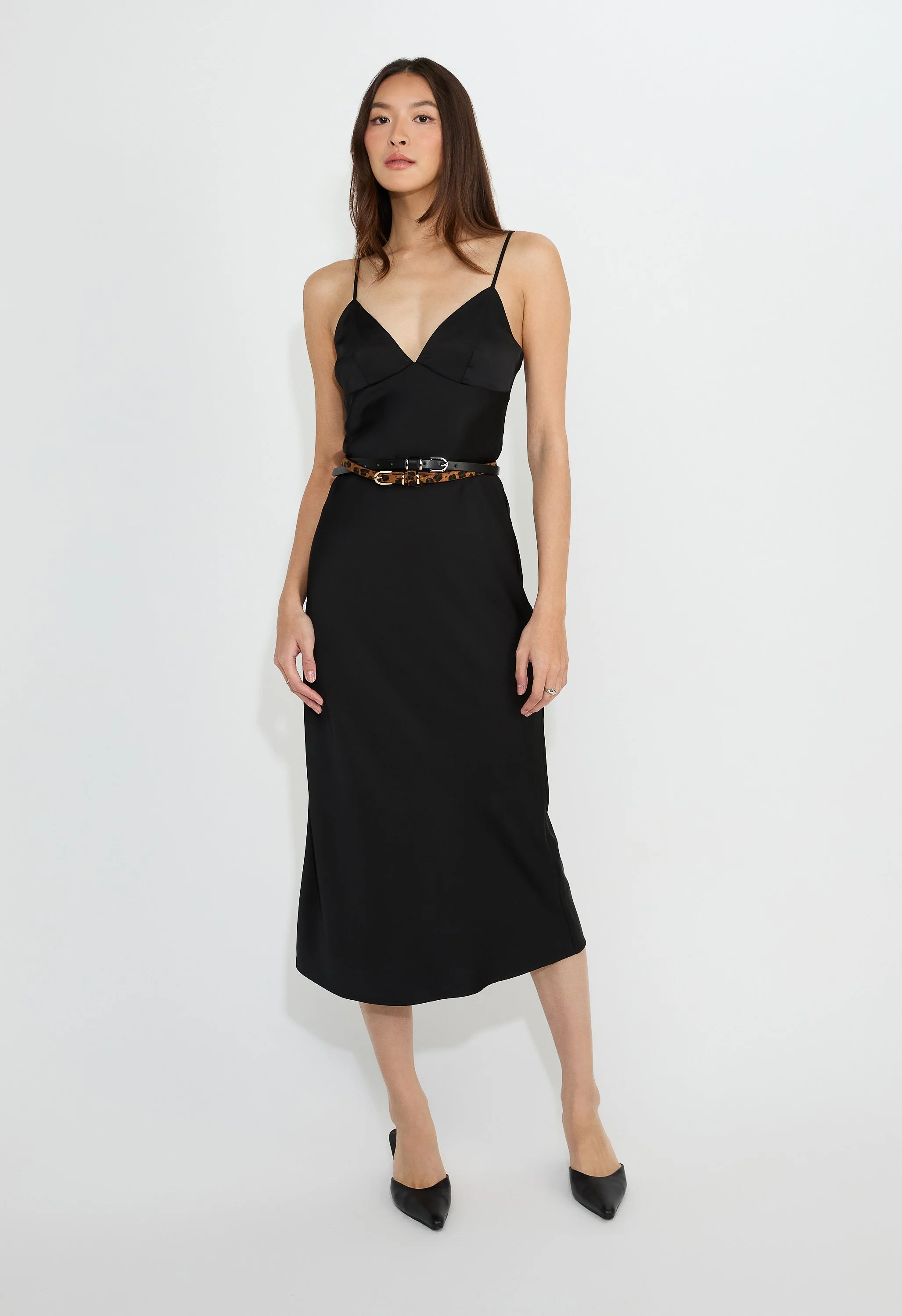 Woman wearing a black spaghetti strap dress with a purple and orange animal print belt, black pointed shoes, standing against a white background.