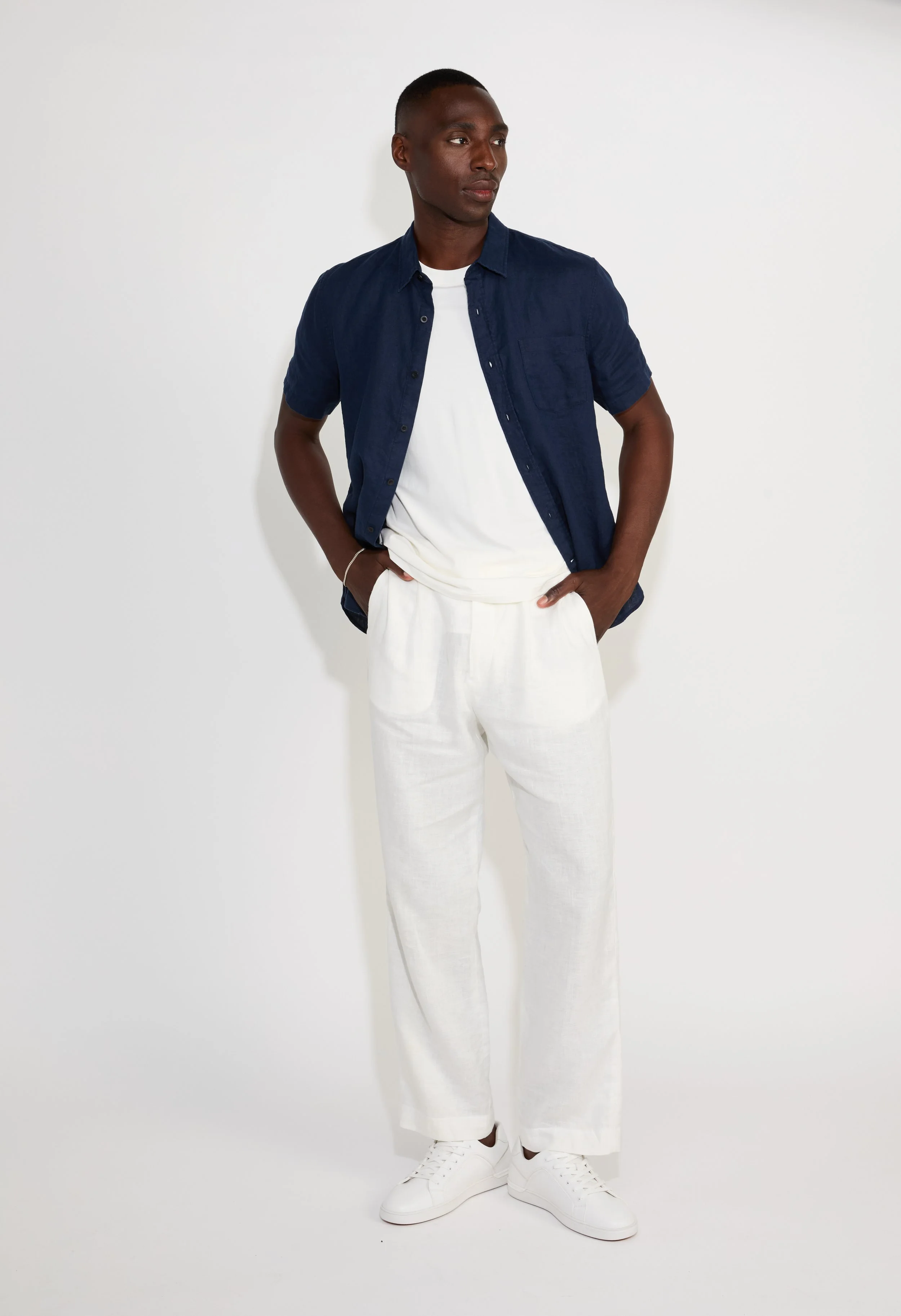 A young man with dark skin and short hair stands against a plain white background. He is dressed in a navy blue casual short-sleeve shirt over a white T-shirt, paired with loose-fitting white pants and white sneakers. His hands are in his pockets and