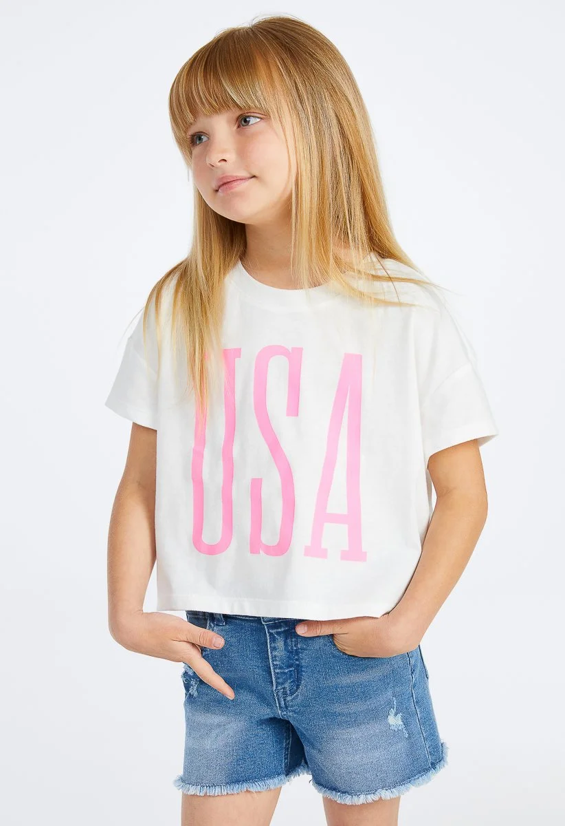 A young girl with long blonde hair wearing a white t-shirt with pink letters spelling 'USA' and denim shorts.