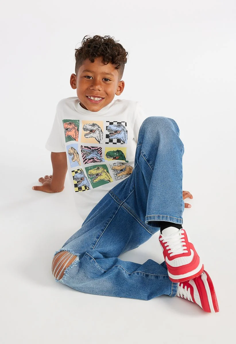 A young boy with curly hair sitting on the floor against a white background. He is wearing a white T-shirt with colorful dinosaur prints, ripped jeans, and red and white sneakers. He is smiling and looking at the camera.