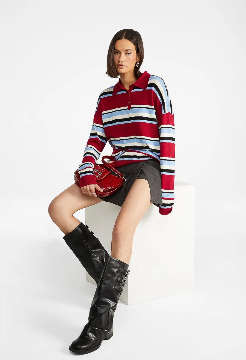Woman in striped oversized polo shirt, miniskirt, and tall black boots, sitting on a white block holding a red handbag against a plain white background.