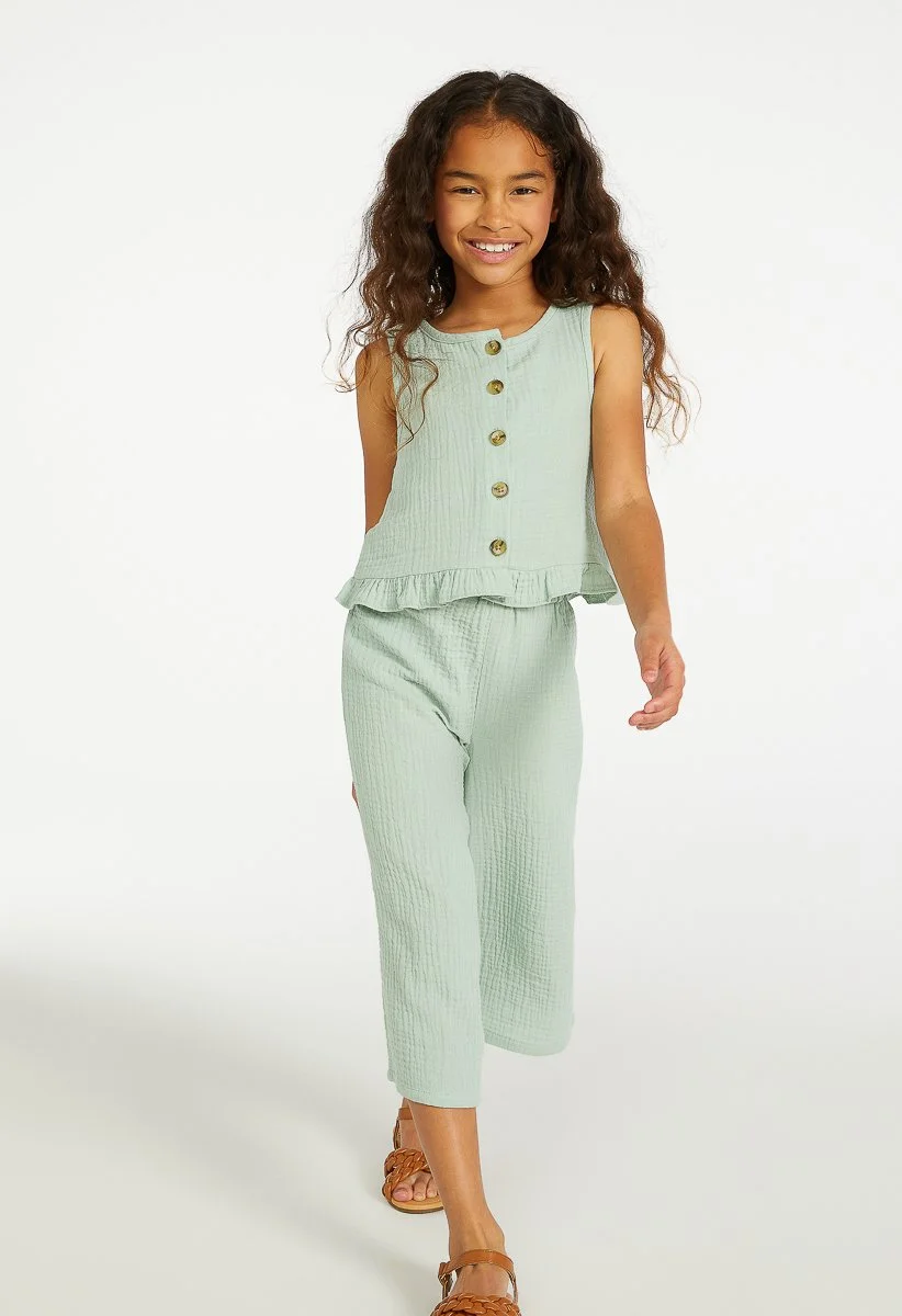 Young girl with long curly hair smiling, wearing a sleeveless light green outfit with buttons and ruffle details, walking barefoot on a white background.