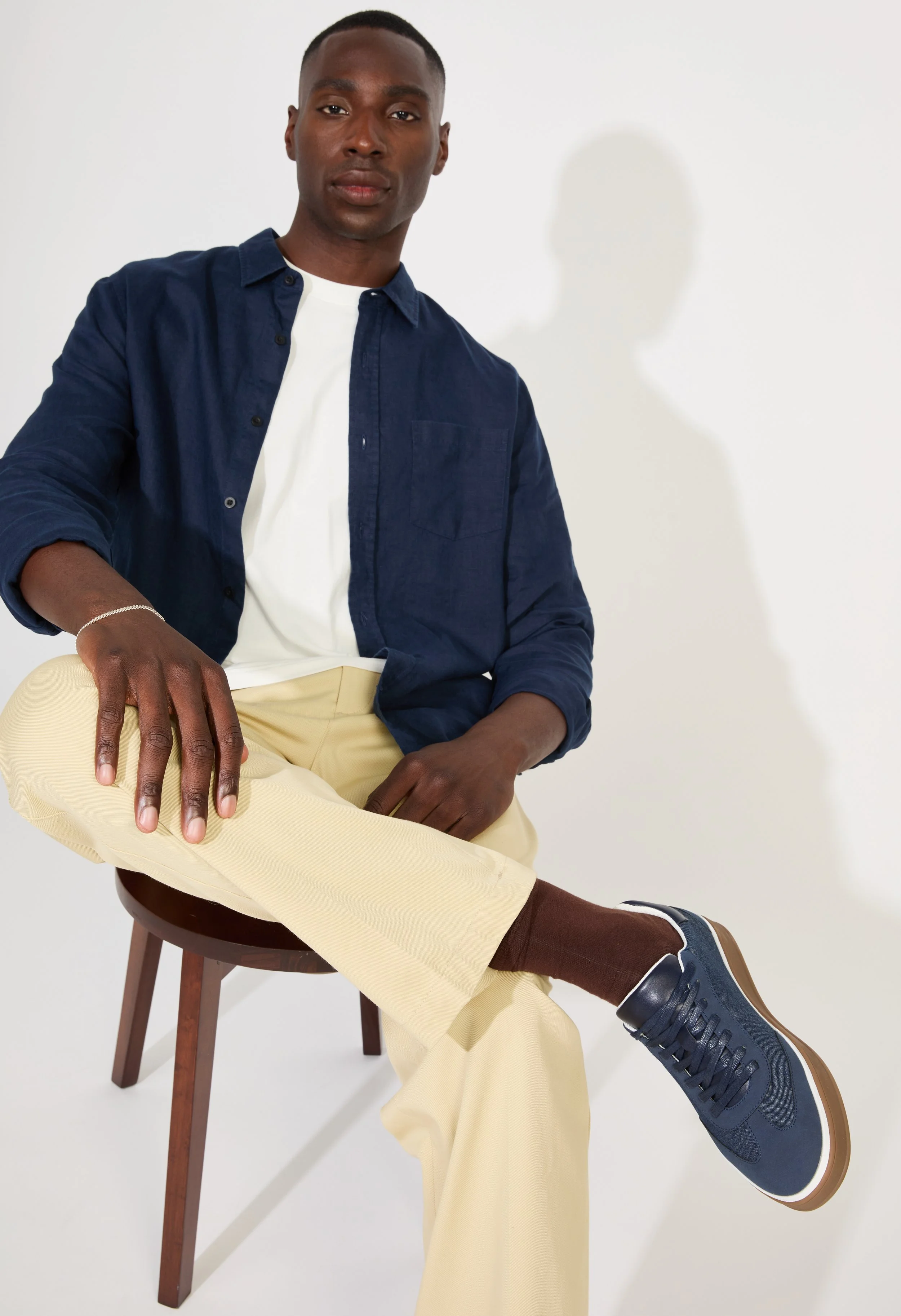 A young man sitting on a wooden chair, dressed in a dark blue button-up shirt, white T-shirt, beige pants, brown socks, and blue sneakers, with one hand resting on his knee and the other on the chair, against a plain white background.