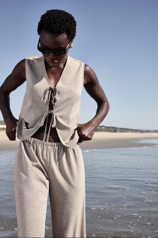 Woman in beige sleeveless top and matching pants standing on the beach with water and sand in the background, looking down.