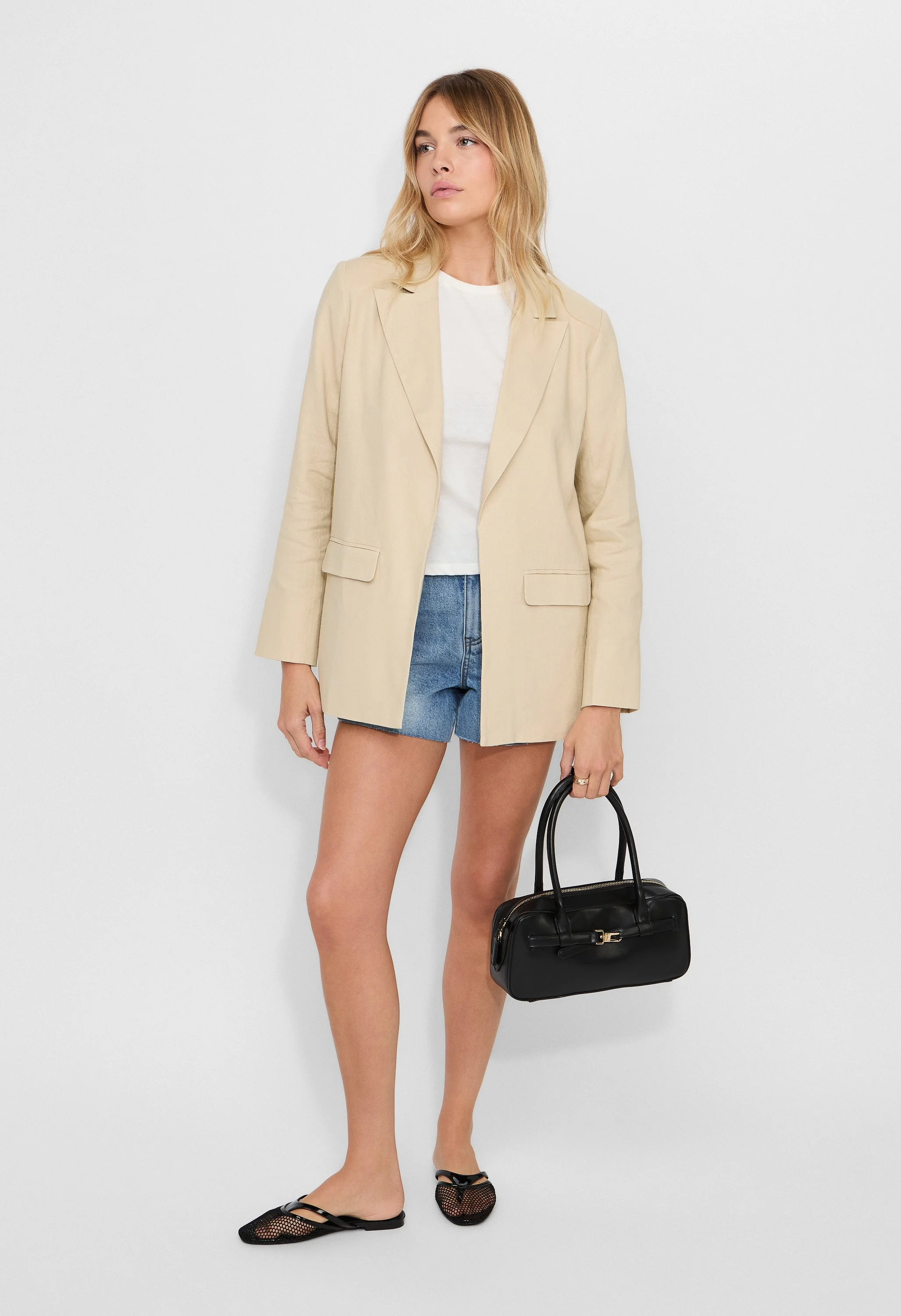 A woman with blonde hair wearing a beige blazer, white t-shirt, denim shorts, black mesh flats, and holding a black handbag against a white background.