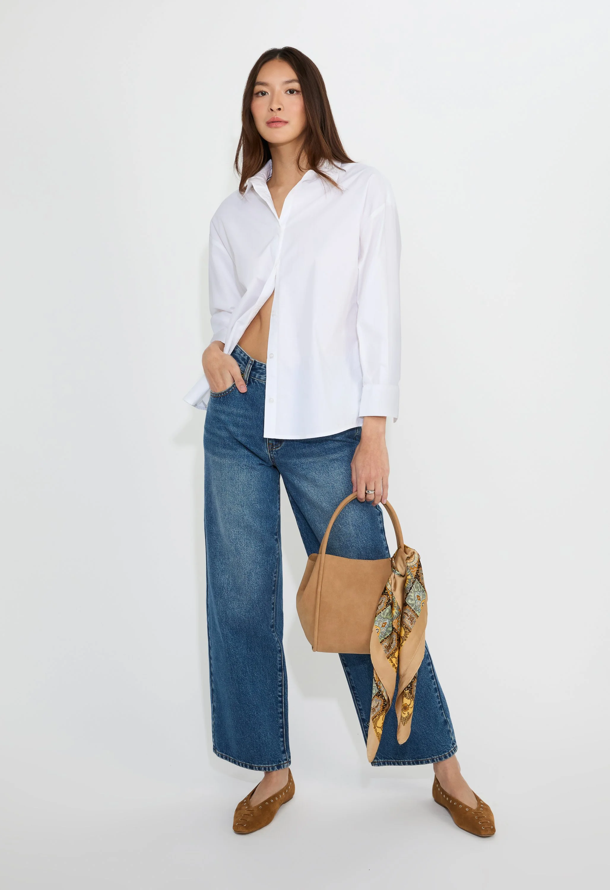 A woman with long brown hair wearing a white button-up shirt, blue wide-leg jeans, brown flats, and holding a tan handbag with a patterned scarf tied to its handle against a plain white background.