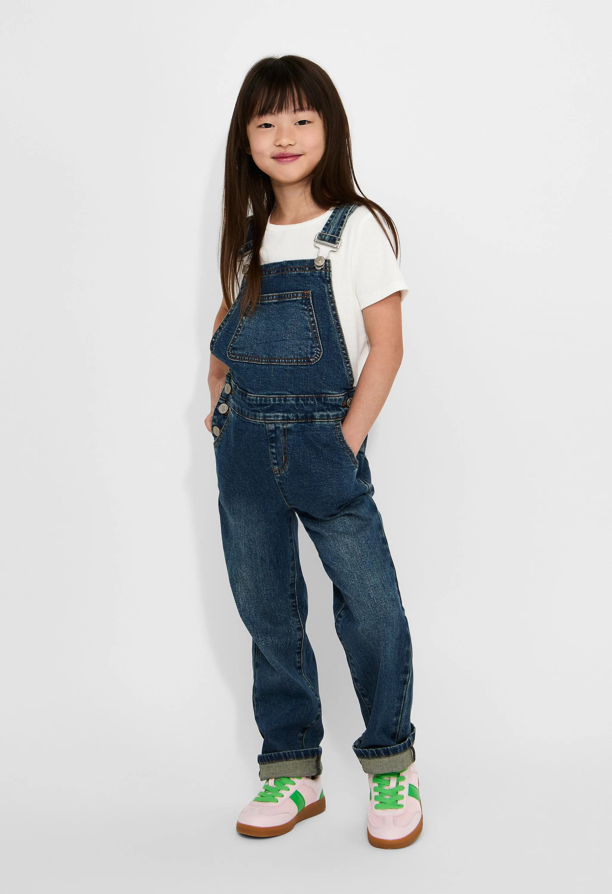 A young girl with long brown hair smiling, wearing a white t-shirt under denim overalls, rolled-up jeans, and colorful sneakers against a plain white background.