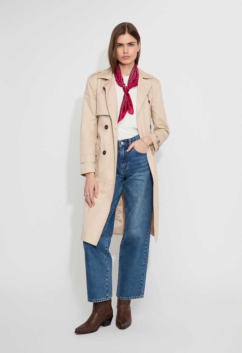 A woman wearing a beige trench coat over a white shirt, blue jeans, brown ankle boots, and a red scarf.