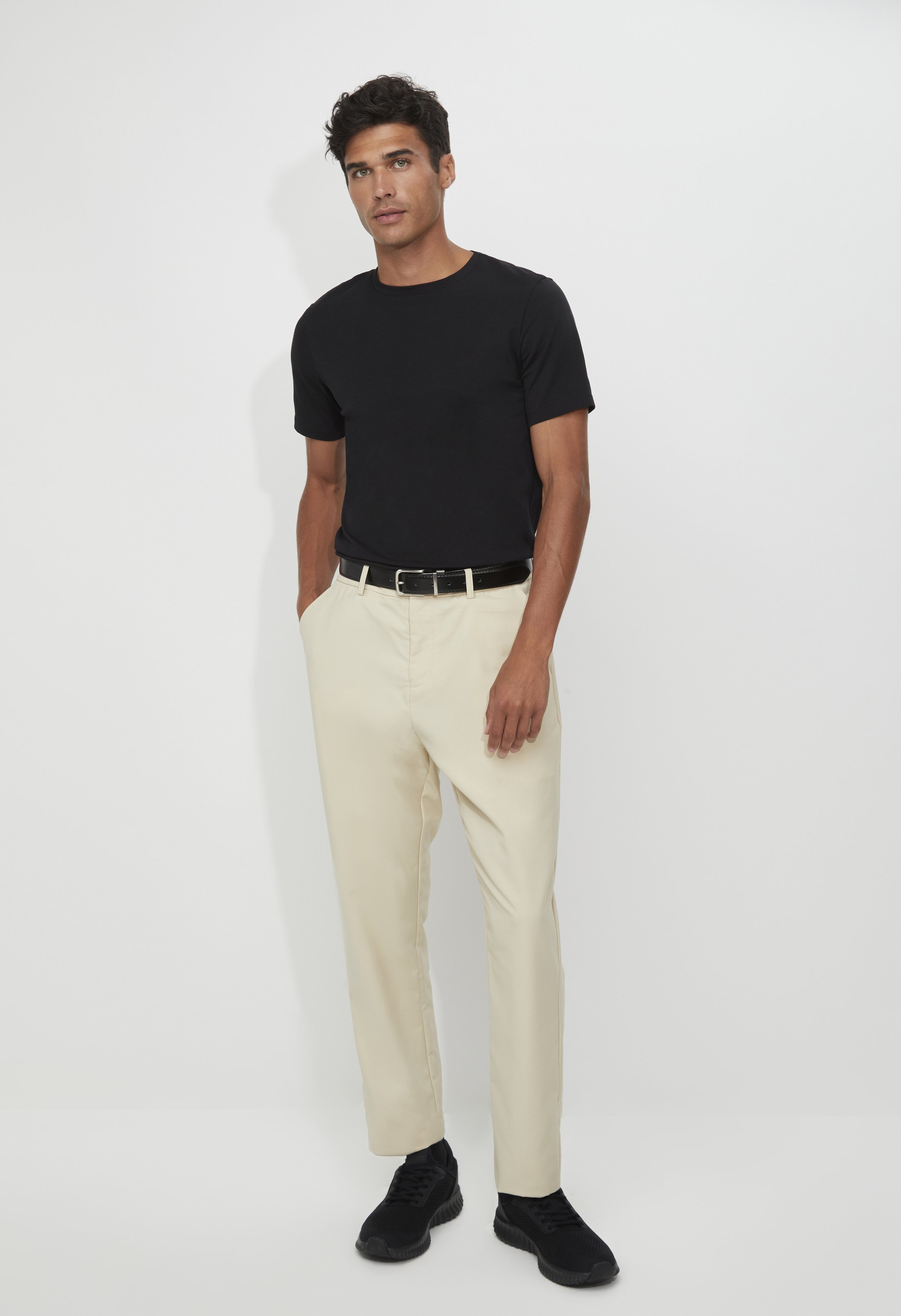 A young man stands against a plain white background wearing a fitted black t-shirt, light-colored trousers, a black belt, and black sneakers.