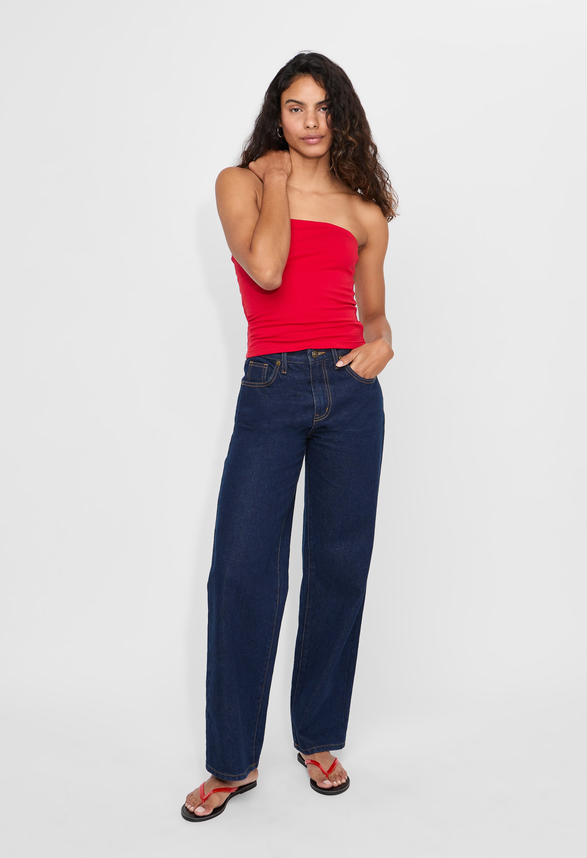 A woman with curly brown hair wearing a strapless red top, dark blue wide-legged jeans, and black flip-flops with red straps. She is standing against a plain white background in an e-commerce studio.