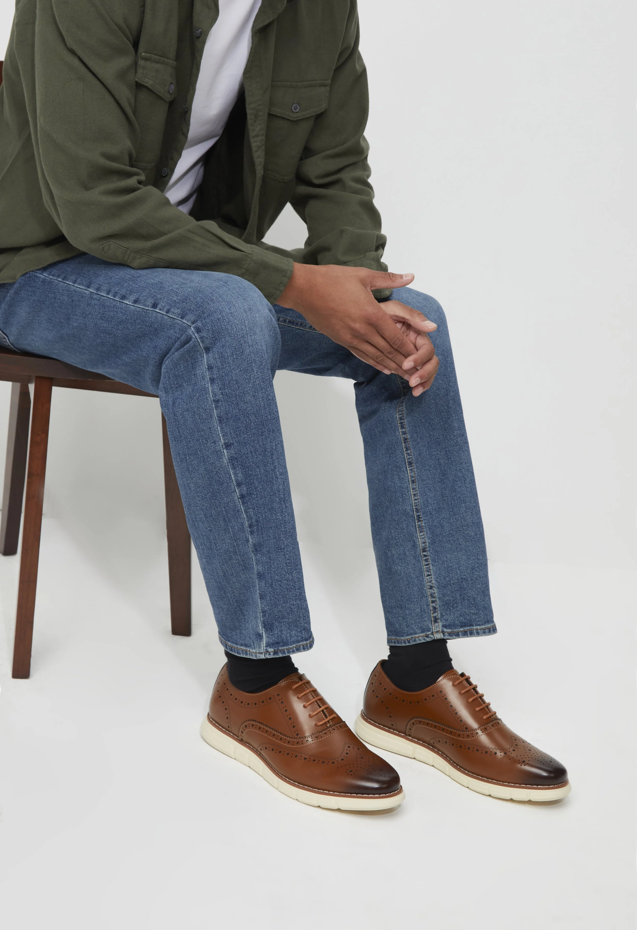 A person sitting on a wooden chair, wearing a green jacket, jeans, black socks, and brown leather shoes with white soles, with hands resting on their lap.