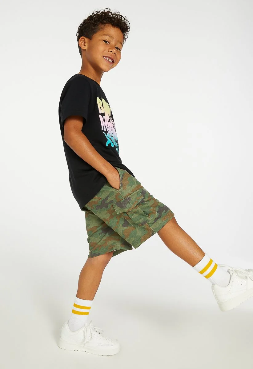 A young boy in a black graphic T-shirt, camouflage cargo shorts, white sneakers, and yellow-striped white socks is standing on one leg, smiling and leaning slightly to the side against a plain white background.
