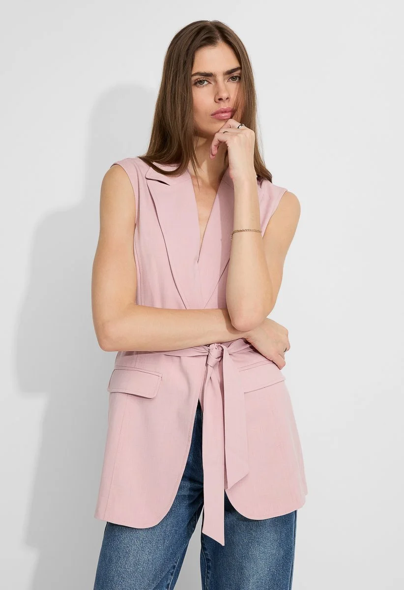 A woman with long brown hair wearing a sleeveless pink blazer with a matching waist tie, posing against a plain white background.