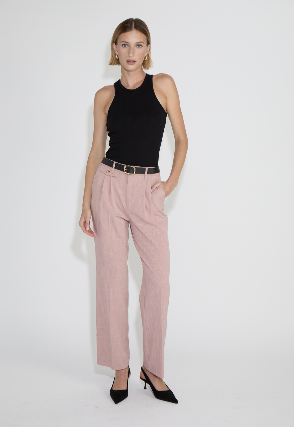 A woman with shoulder-length blonde hair wearing a black sleeveless top, pink trousers, a black belt, and black heels, standing against a white background in an e-commerce studio.