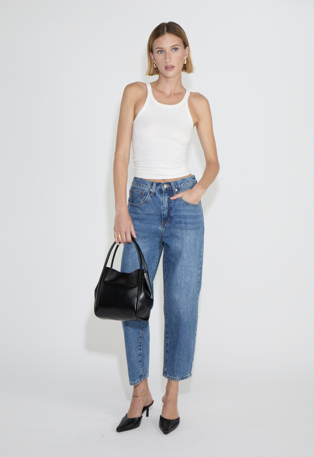 A woman wearing a white tank top, blue jeans, black heels, holding a black handbag, standing against a white background in an e-commerce studio.