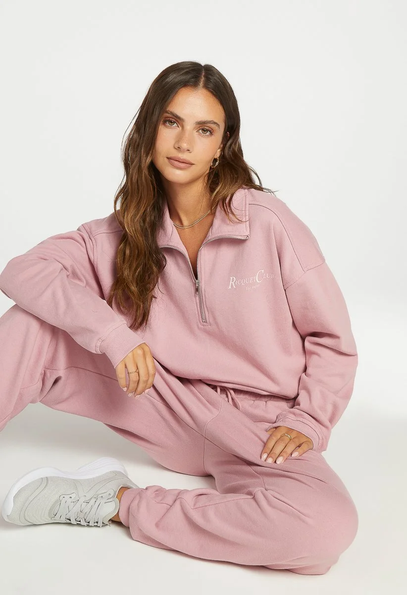 A woman with long brown hair sitting on the floor against a plain white background, dressed in a light pink sweatshirt and matching sweatpants, wearing white sneakers and minimal jewelry.