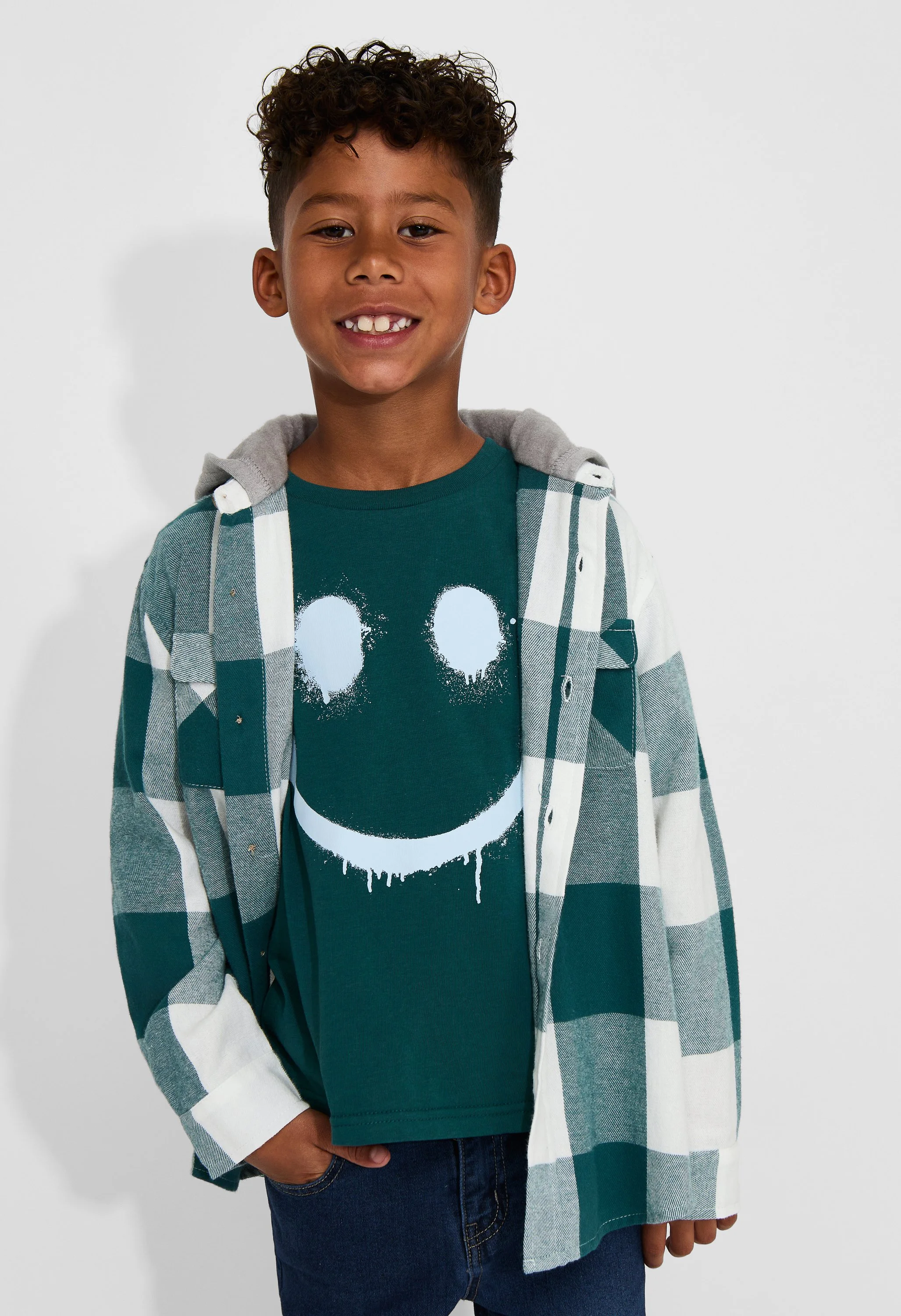 A young boy with curly hair is smiling. He is wearing a teal T-shirt with a white skull graphic, a checkered button-up shirt, and dark jeans, standing against a plain white background.