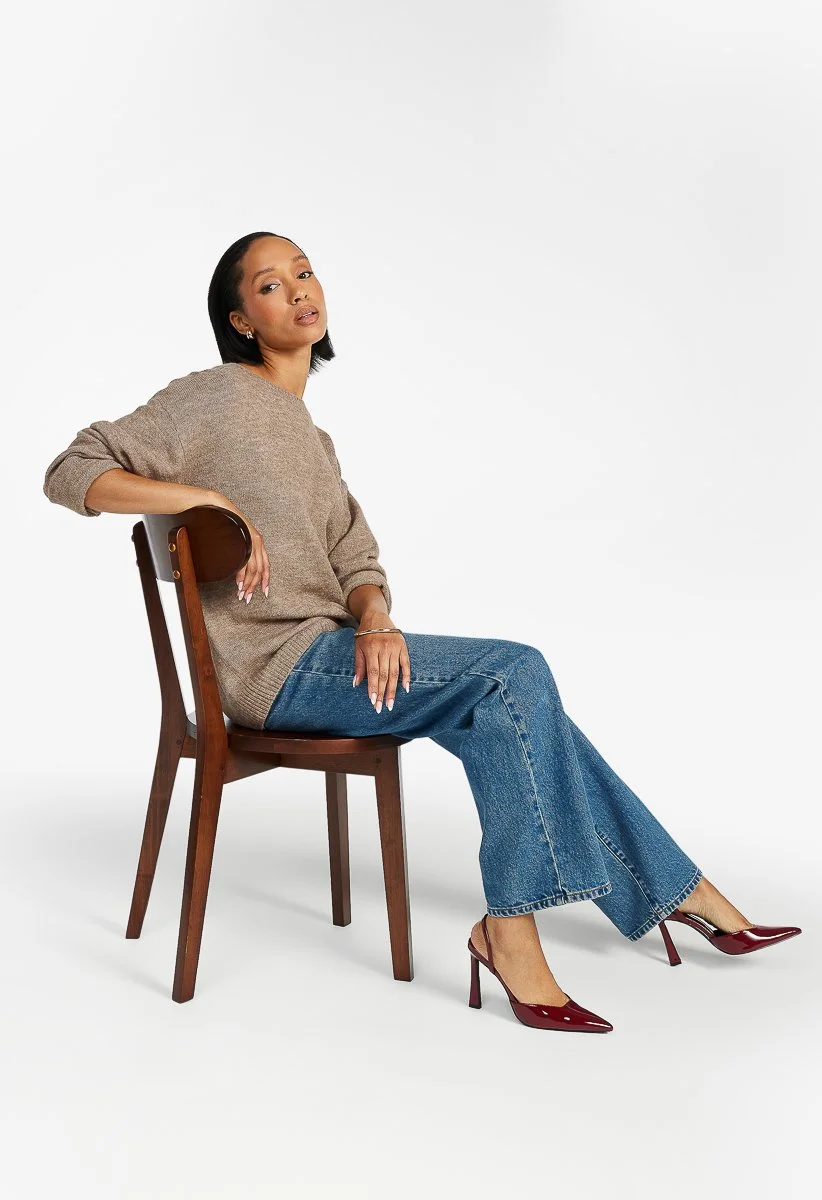 A woman sitting sideways on a wooden chair with one arm resting on the backrest, wearing a beige sweater, blue jeans, and burgundy high heels, against a plain white background in an e-commerce studio.