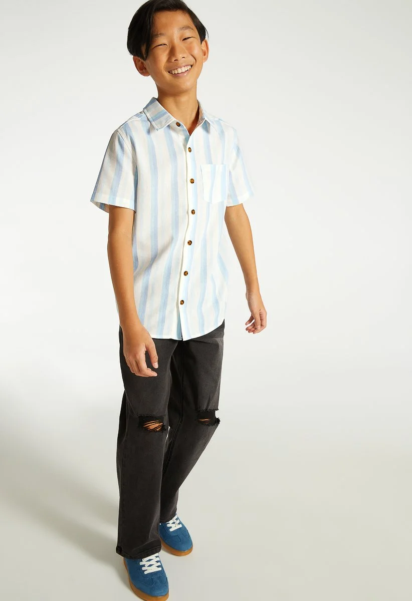 A smiling boy wearing a short sleeve, button-up shirt with light blue and white vertical stripes, black ripped jeans, and blue sneakers, standing against a plain light background.