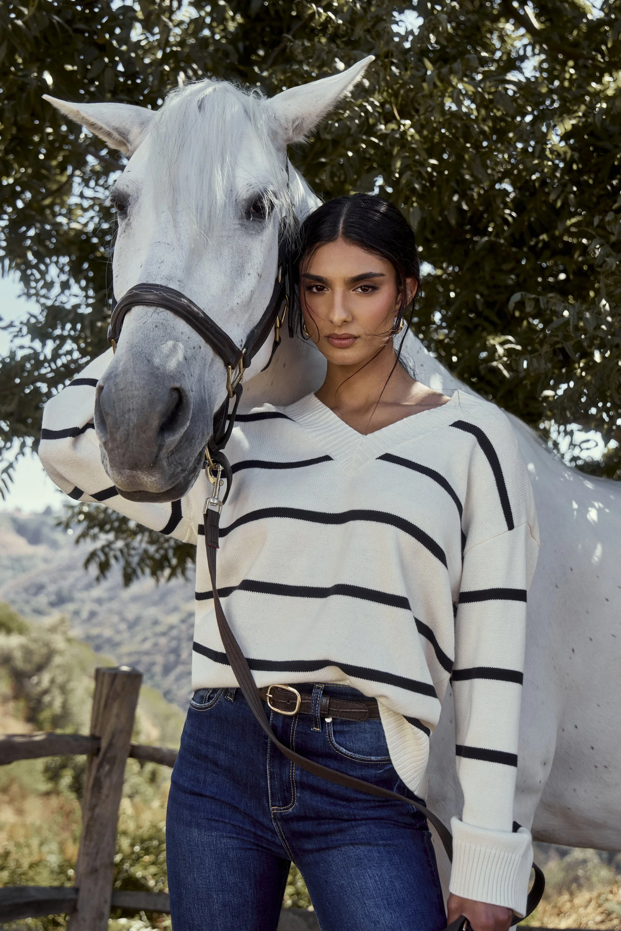 A woman with dark hair in a ponytail, wearing a white sweater with black stripes, is standing outdoors next to a white horse with a black bridle. The background features trees and a scenic landscape.